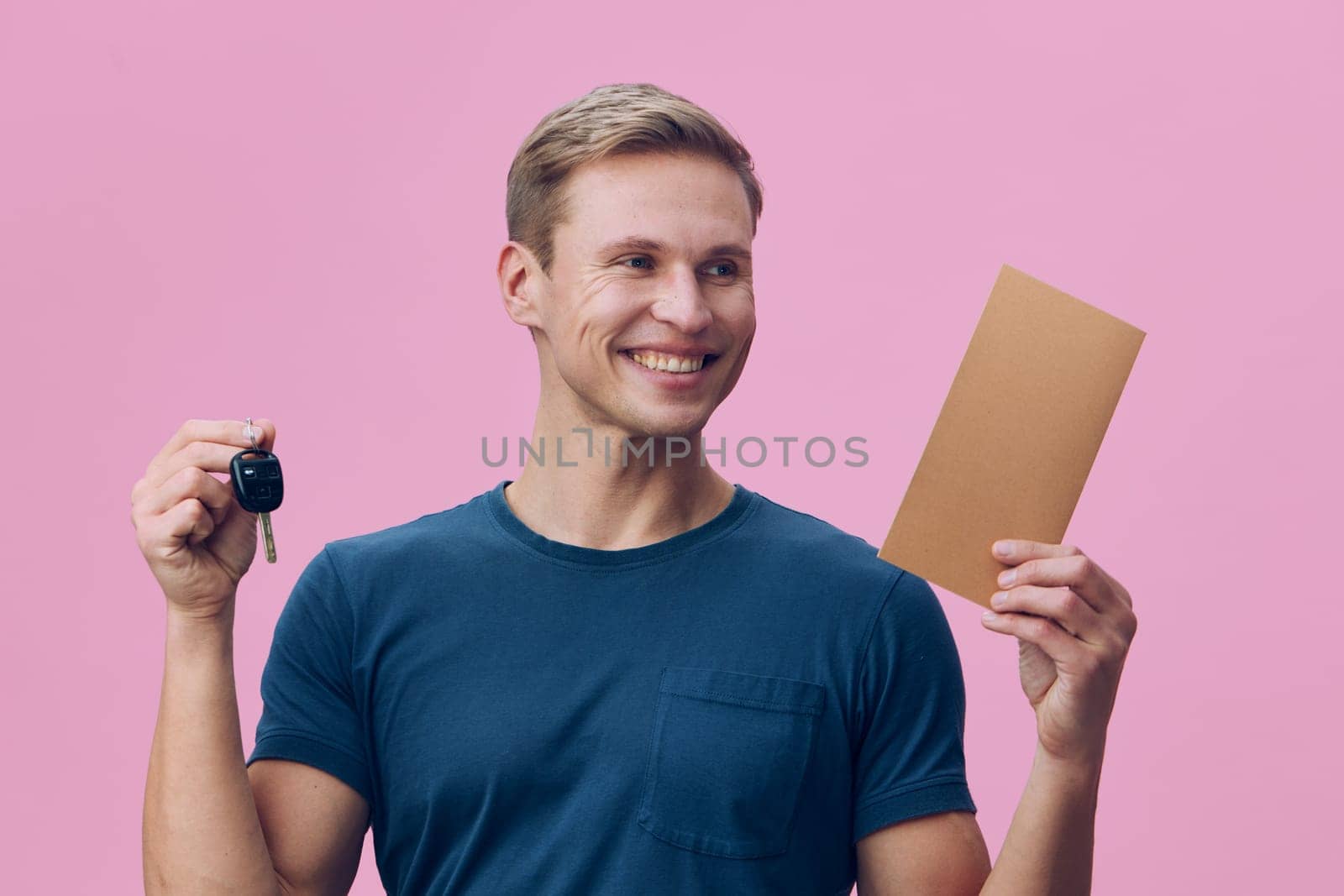 Happy man with short blond hair presenting car key and cardboard box, smiling confidently on pink background. Casual attire, joyful mood, studio portrait, celebration, success, friendly atmosphere. Concept: achievement and new beginnings.