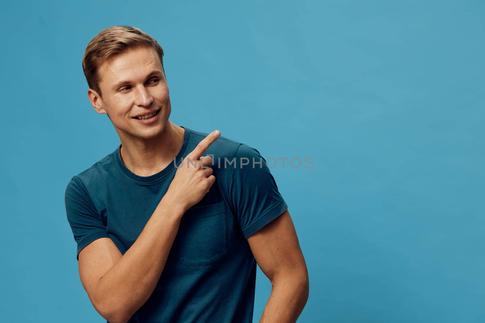 Smiling young caucasian man wearing blue t-shirt looking and pointing finger to side standing isolated on plain blue background with casual style. Bright expression showing positive emotion. Concept: digital nomad lifestyle