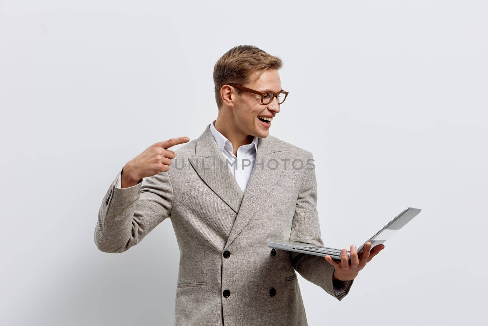 Happy man in stylish blazer and glasses, holding a laptop and gesturing towards his head, bright expression, modern office environment, positive mood, work achievement, technology device, professional lifestyle.