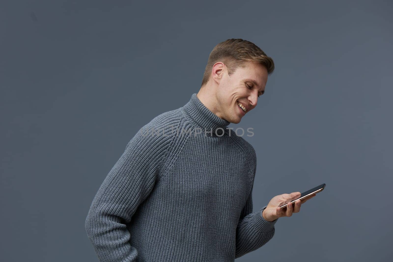 Happy man with short hair in cozy gray knit sweater looking at smartphone with smile against simple gray backdrop. Concept: enjoyment of digital communication, relaxed lifestyle, modern technology.