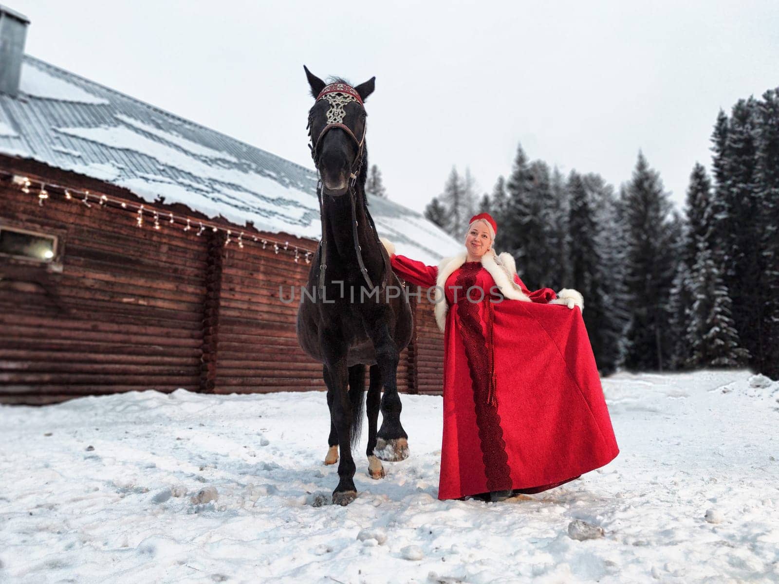 Girl in a colorful national dress stands by a horse in the snowy landscape, celebrating Maslenitsa.