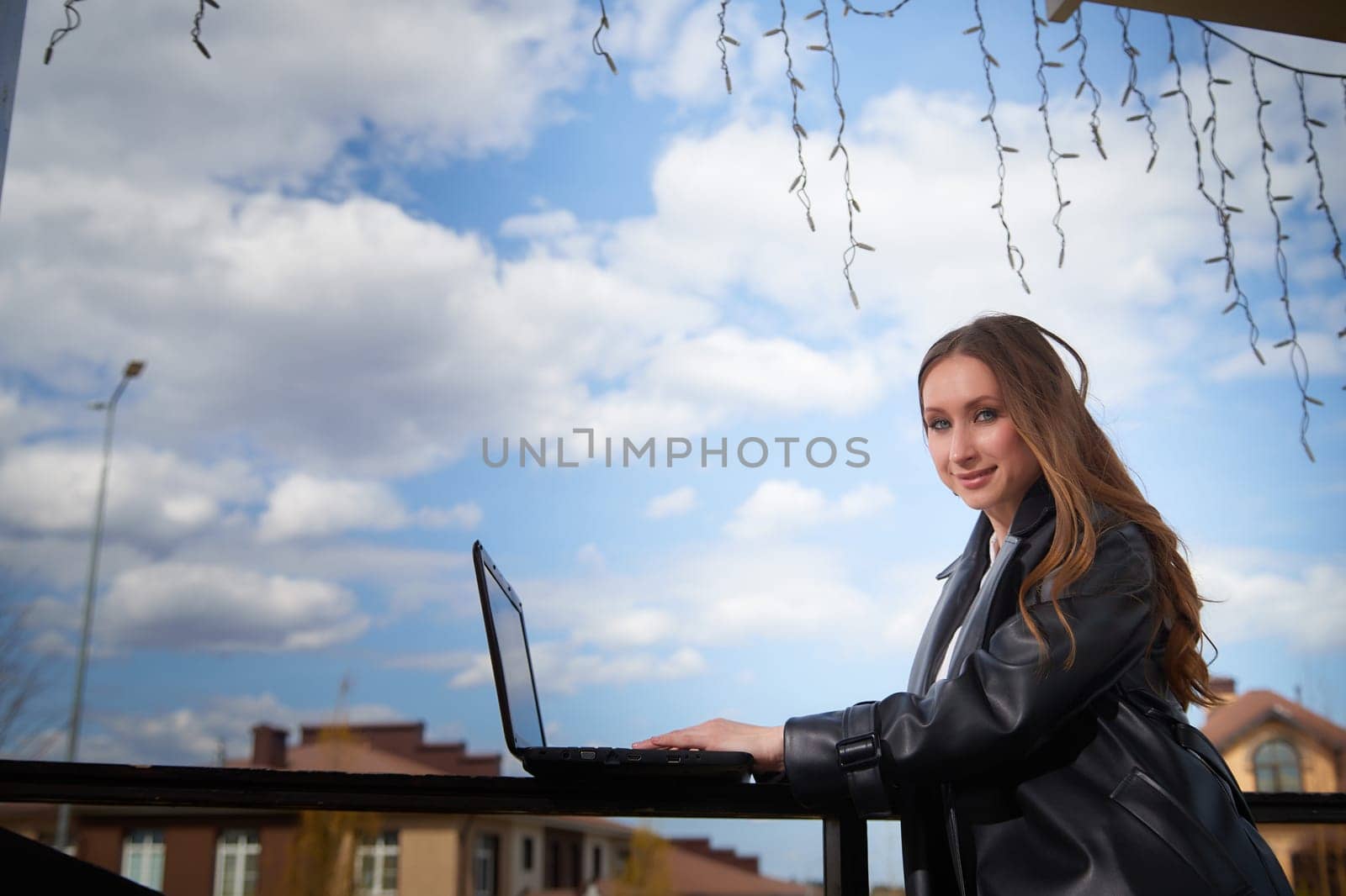 A young woman sits at a table using a laptop while enjoying a beautiful day outdoors under a partly cloudy sky.