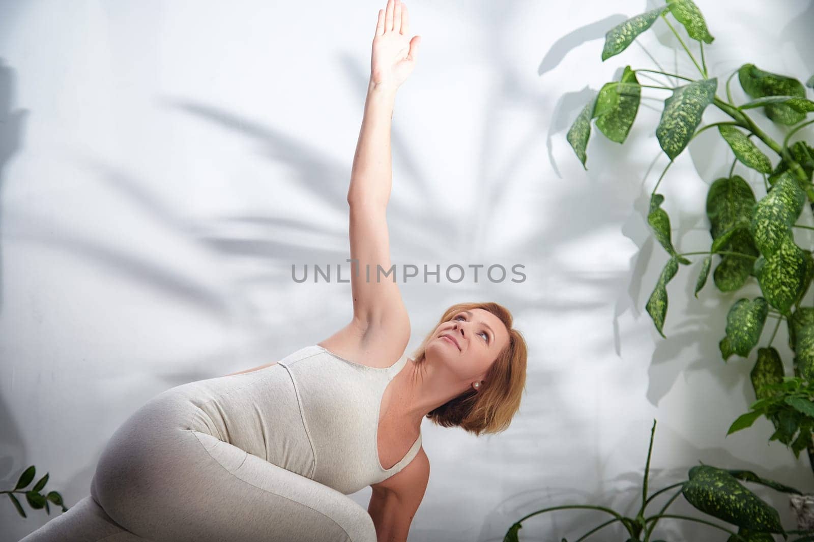 A woman performs a yoga pose promoting flexibility and mindfulness in a peaceful indoor environment enriched with greenery.