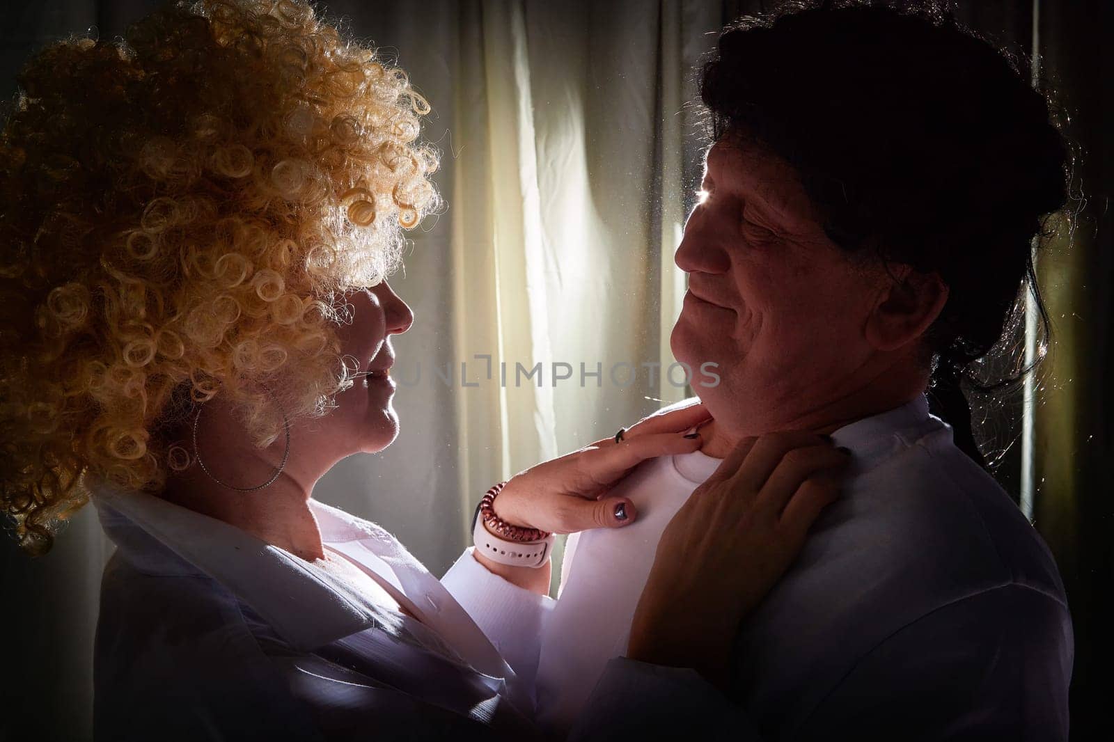 A middle-aged couple engages in a theatrical photoshoot indoors, wearing vintage wigs and showcasing their love in a warm, personal atmosphere.