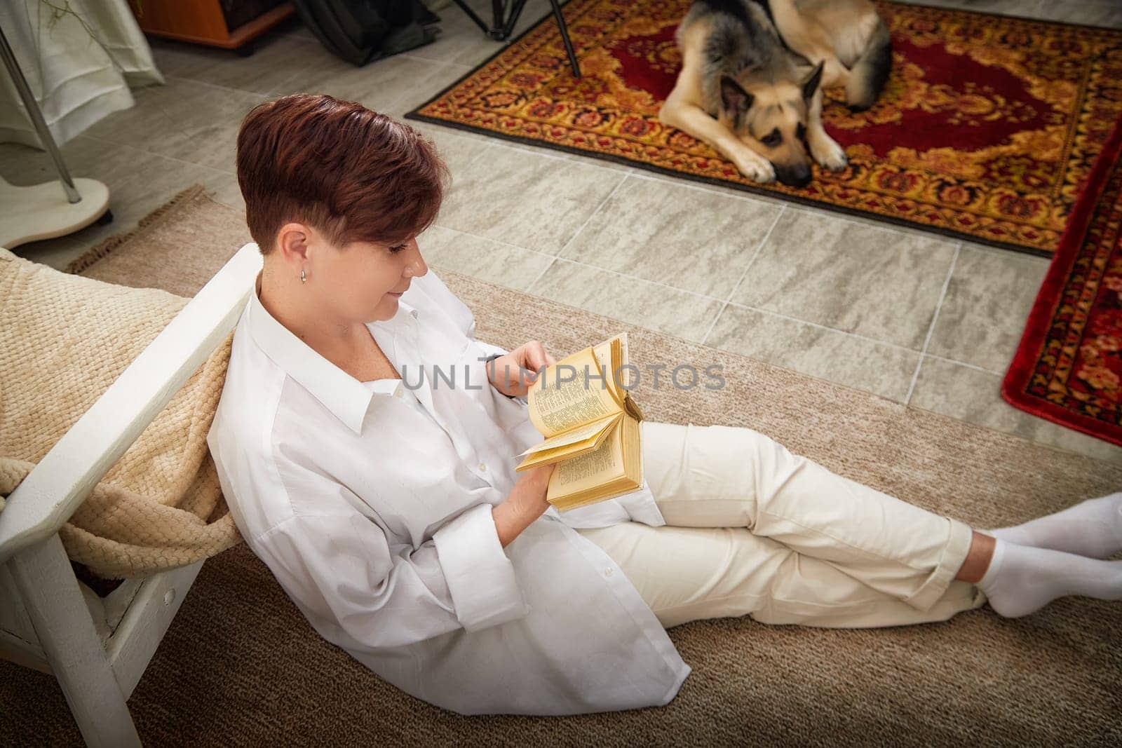 Middle-aged woman sits on the floor reading a book, enjoying a peaceful moment with her dog in a cozy room.