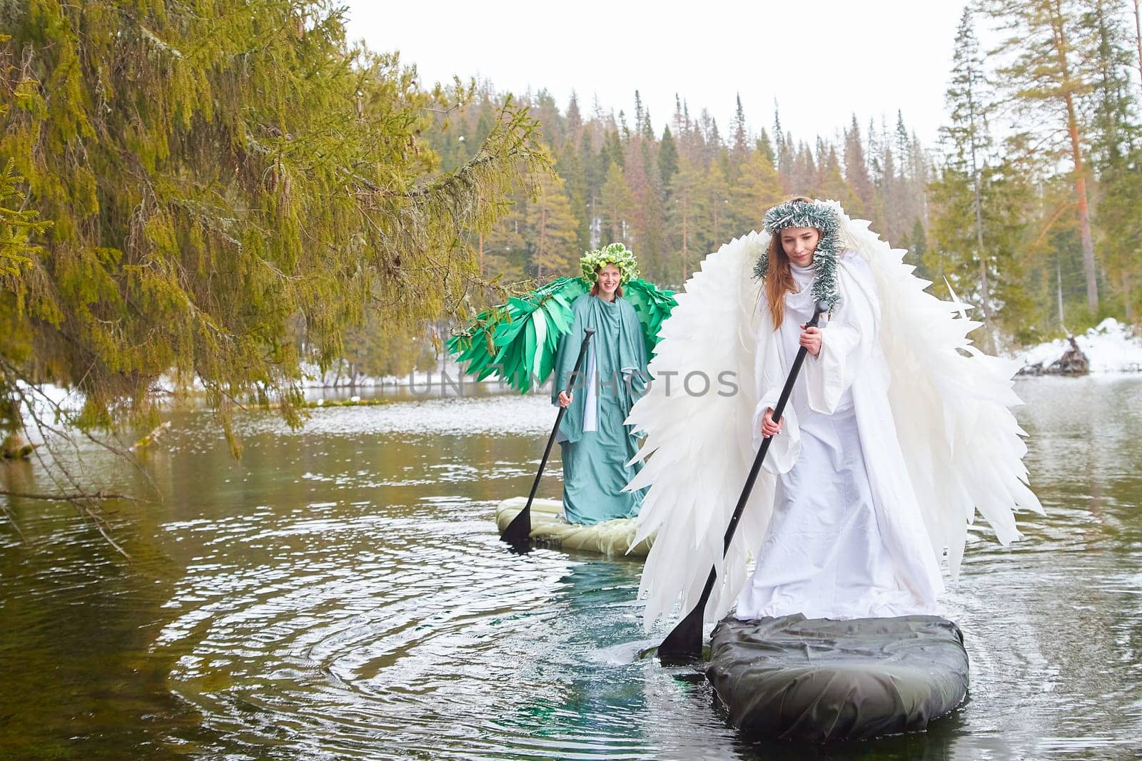 Two girls dressed in flowing white and green garments paddle across a tranquil lake, embodying the transition between winter and spring.