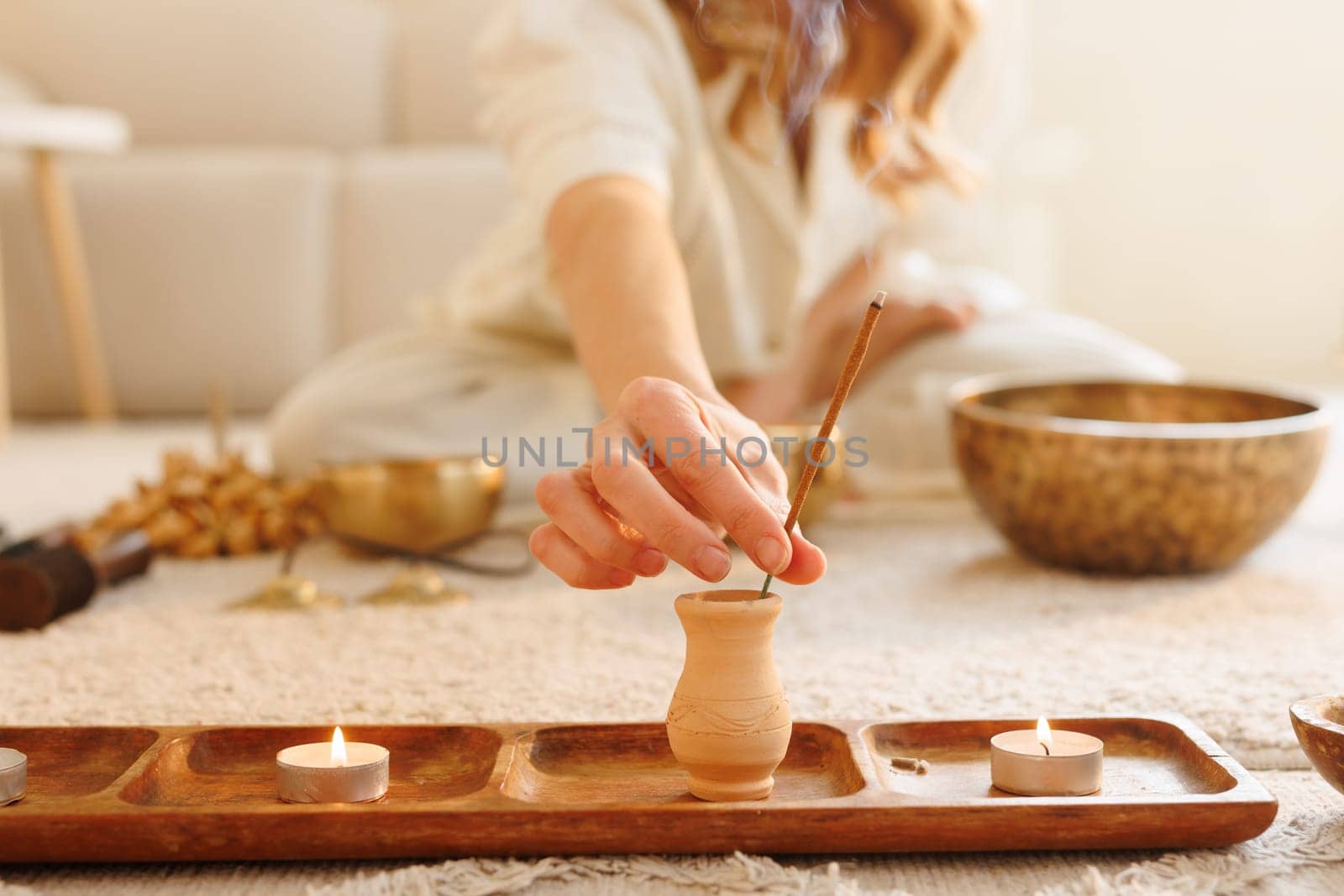 Woman lighting incense stick in cozy space with candles and Tibetan bowls for meditation and energy cleansing.
