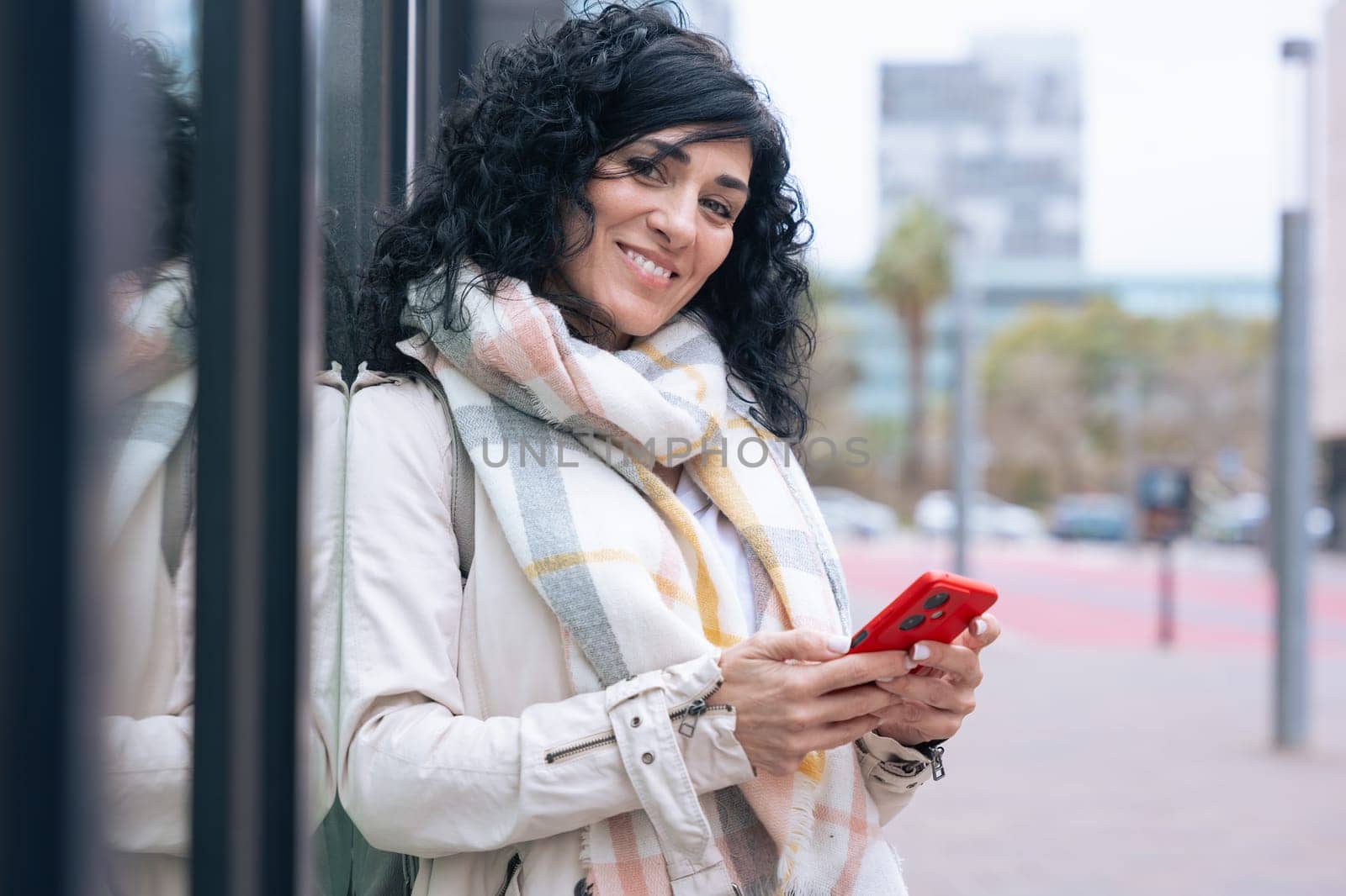 Happy caucasian woman standing outdoors in urban setting,wearing coat and scarf,using her red smartphone.She is smiling and relaxed,enjoying a casual moment on a cold day in the city looking at camera