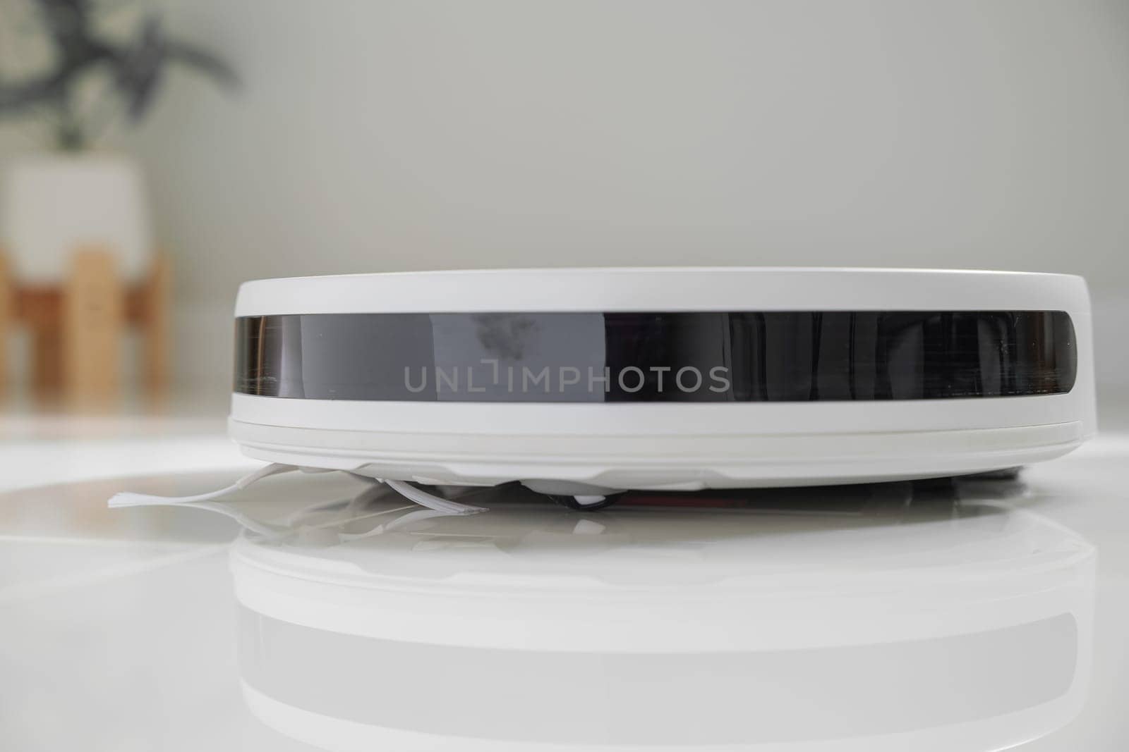 White automatic floor cleaner with side brush on tile flooring, featuring front black sensor strip. Robot vacuum cleaner efficiently cleaning floor, illustrating smart home technology for dust removal