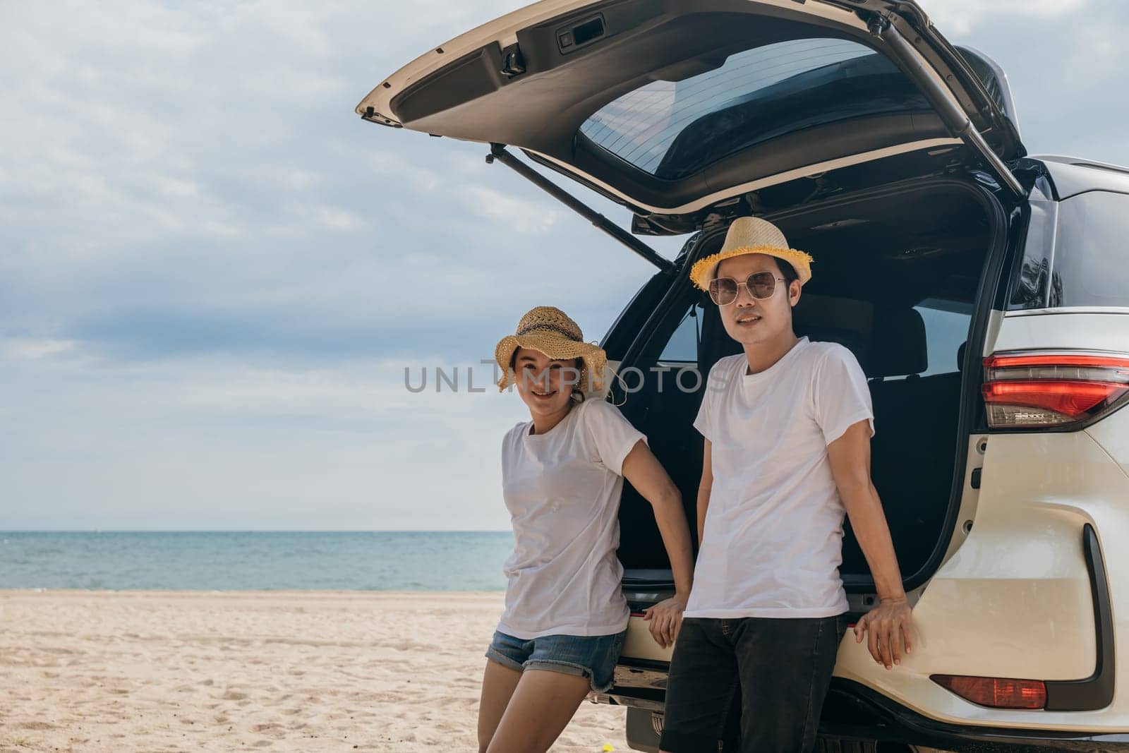 Asian couple is posing in back of a car enjoying beautiful views on the ocean beach. lifestyle man and the woman is wearing hat. Scene is relaxed and carefree, love and travel summer trip concept