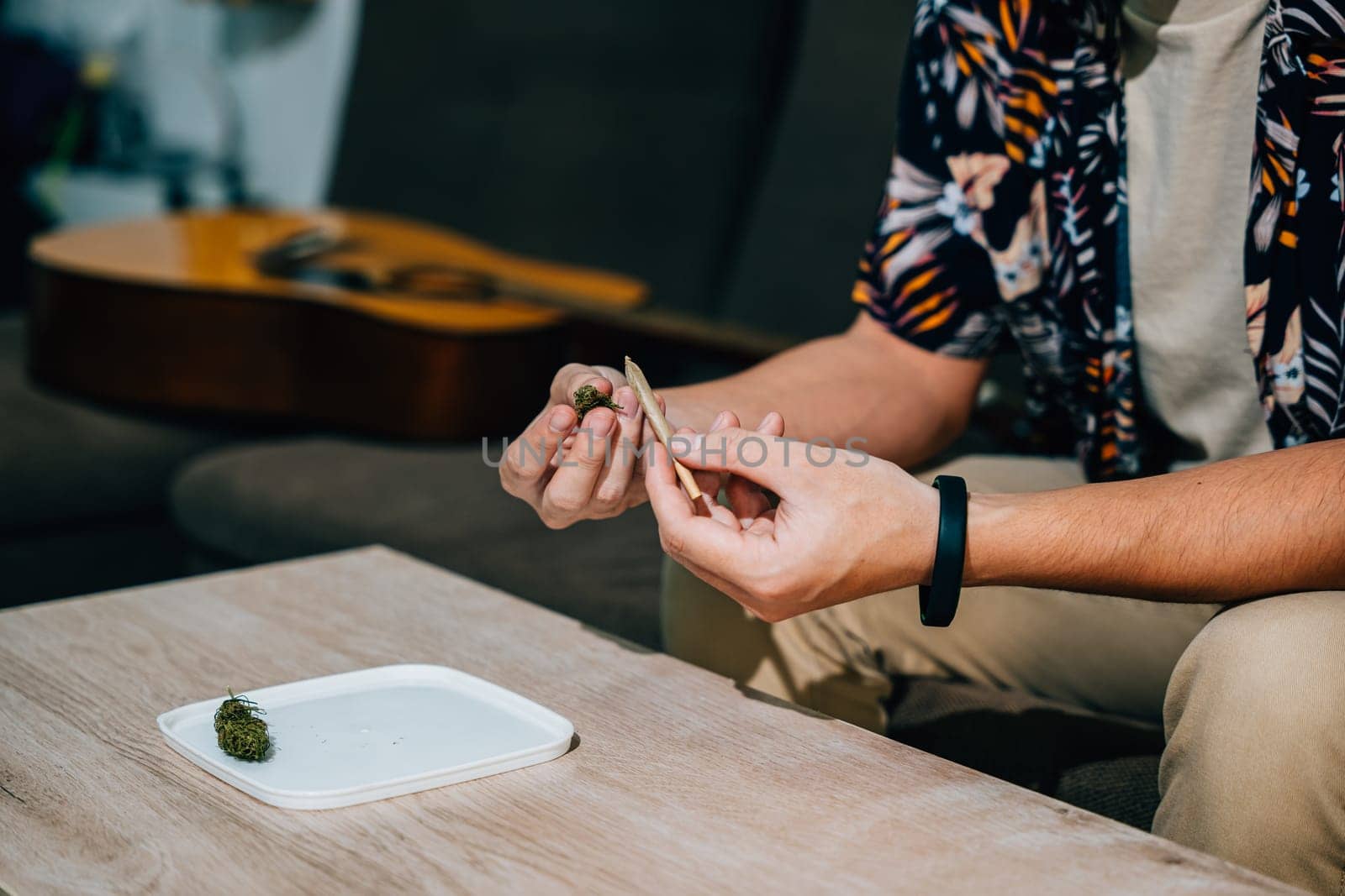 Asian young man sitting on a couch with a cannabis bud in his hand and smoking a joint relaxing time at home. A guitar is in the background