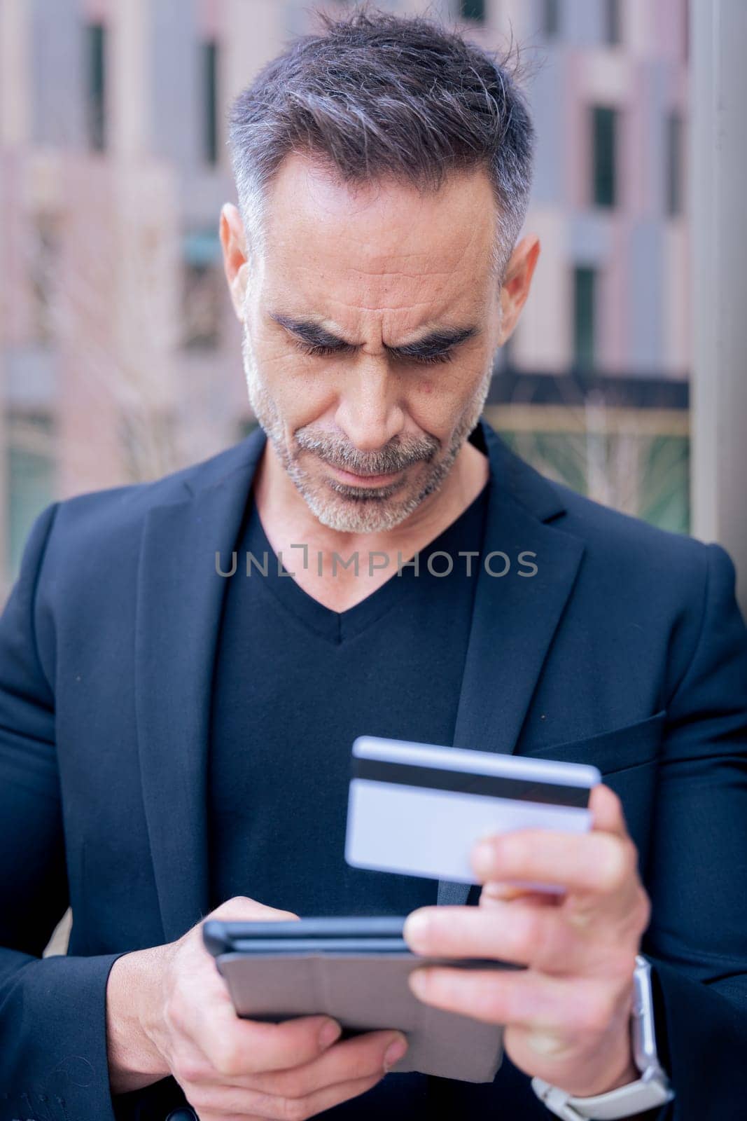 Confident caucasian man with headphones serious while using tablet and holding a credit card outdoors. Concept of online shopping, digital payment, modern lifestyle, and financial technology. Vertical