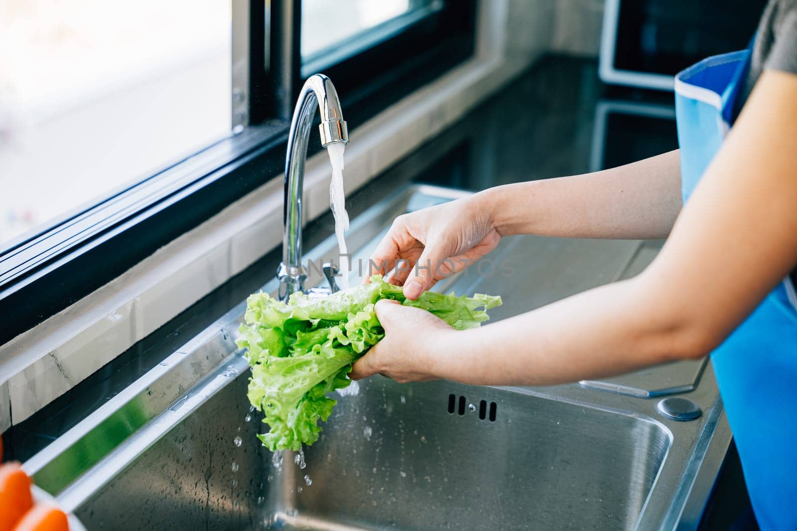 In a modern kitchen a woman's hands wash fresh vegetables under running water at the sink for preparing a vegan salad. Homemade healthy food concept with clean bright leafy greens.