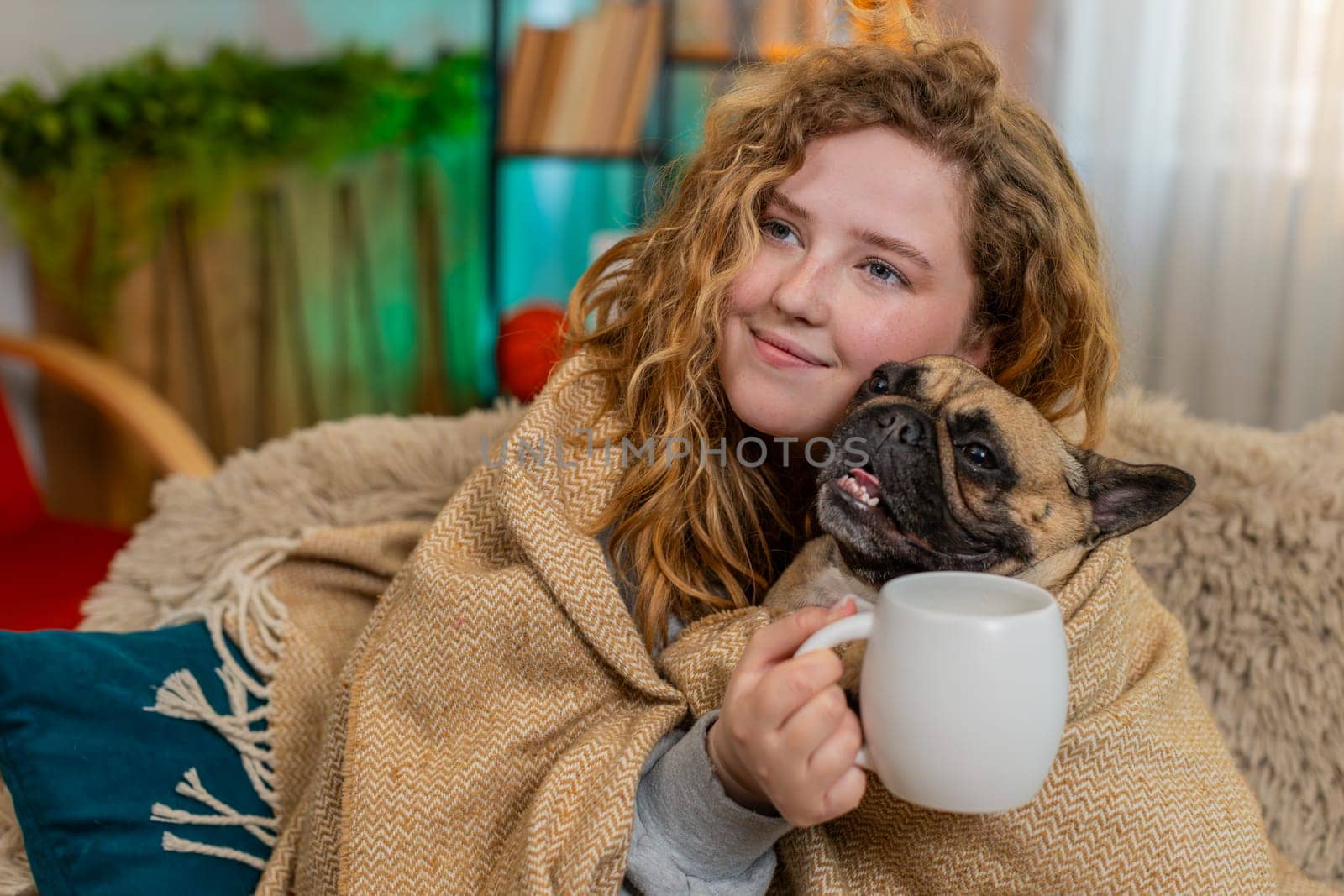 Young Caucasian redhead woman enjoys tea under blanket on home sofa, holding cup with warmth and calm. Pug dog rests beside her under same blanket, sharing comfort and peaceful happy family moment.