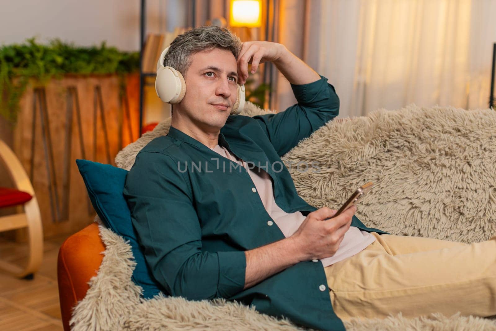 Young man at home lies on sofa with eyes closed and headphones on, listening to audiobook. Caucasian guy looks calm and peaceful, enjoying relaxation and imagination from the comfort of quiet space