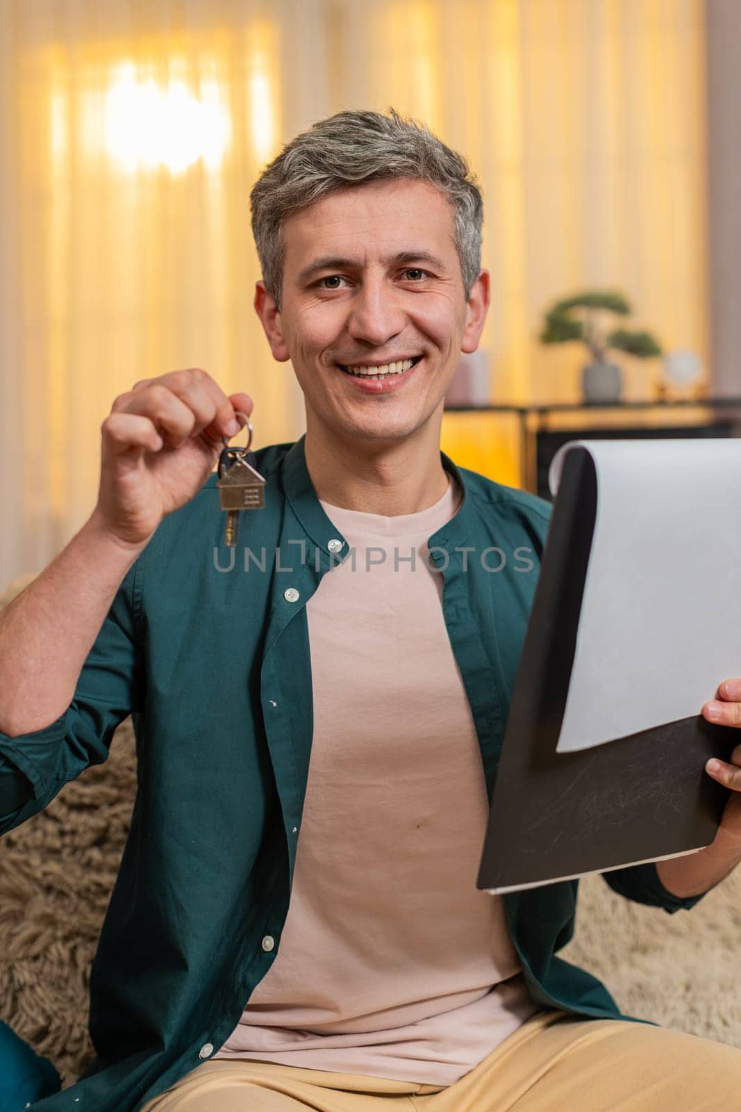 Young man at home smiles while holding keys and property documents in hand, proud and satisfied. Caucasian guy on sofa celebrates apartment deal and feels secure, confident and emotionally uplifted.