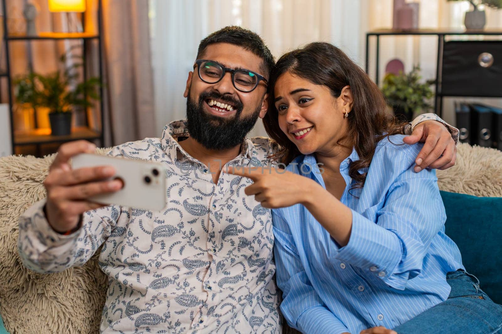 Indian couple man and woman sitting on home sofa, laughing while watching funny social media videos online together on smartphone. Young family enjoying fun moment and digital content indoors with joy