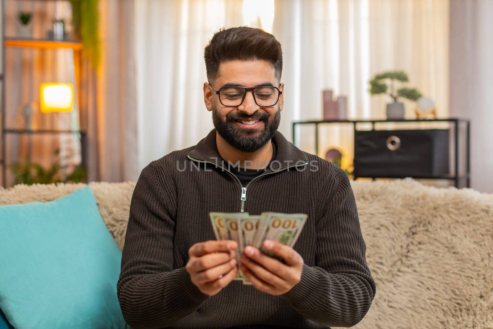 Indian man at home happily counts cash dollars while sitting on sofa, enjoying recent income or lottery win. Arabian guy expresses joy and financial confidence, celebrating success with satisfaction.
