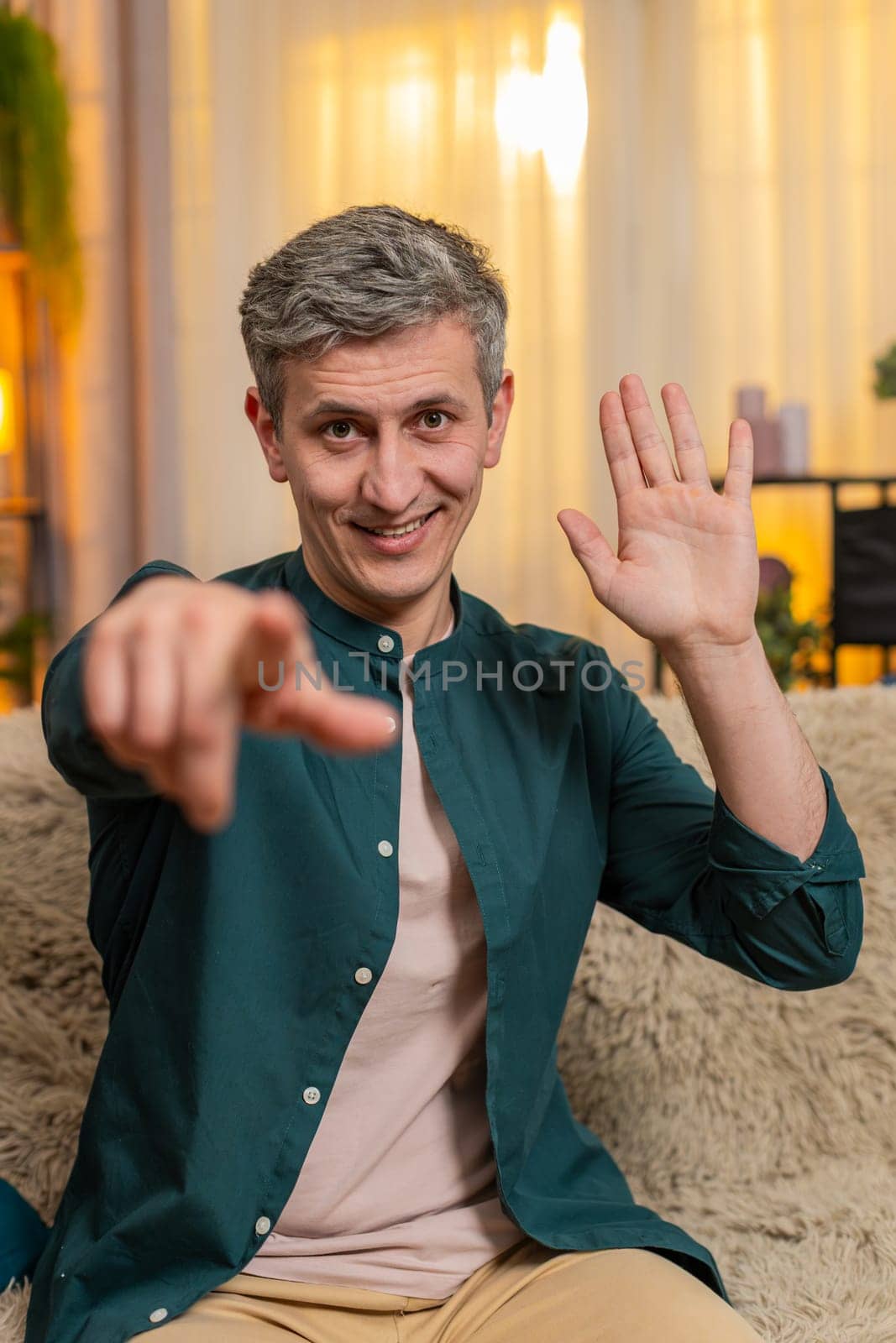 Young happy man at home pointing at camera with smile during video call greeting excitedly. Caucasian guy on sofa shows joy happy energy and enthusiasm, saying hello with expressive friendly attitude