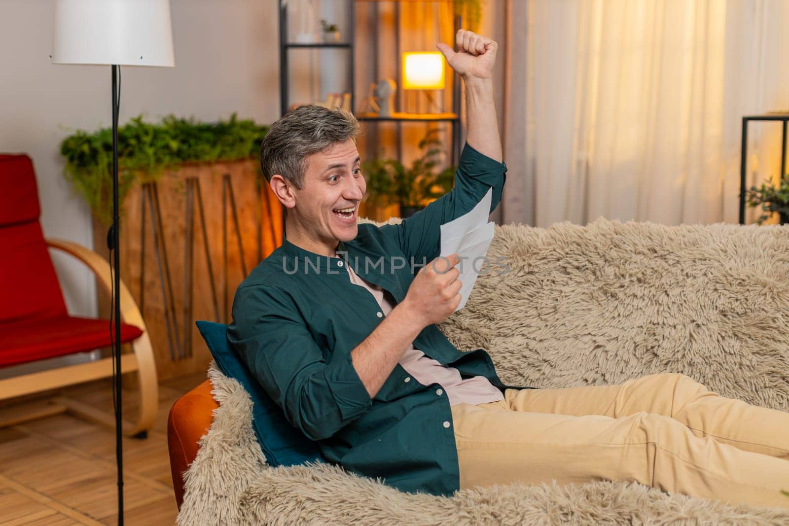 Young happy man at home opens letter and happily celebrates great news, raising fist with joy. Caucasian guy on sofa feels excited about job offer, loan approval or successful school exam result.