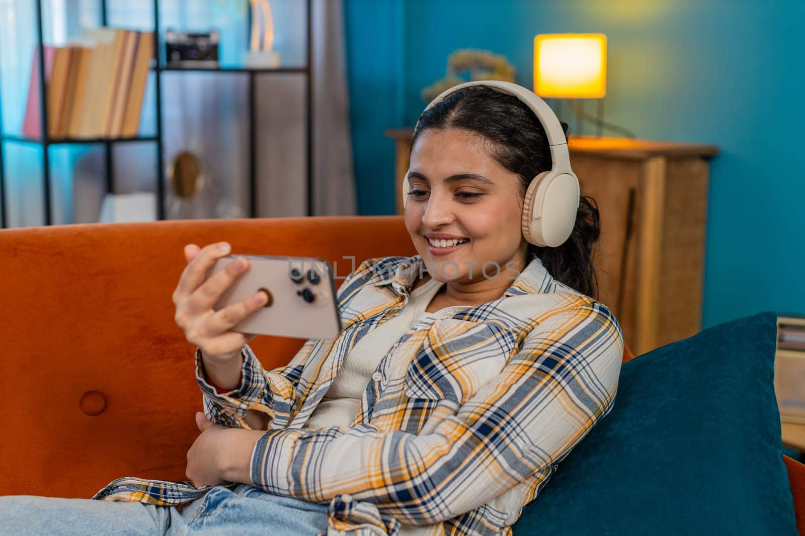 Indian young woman relaxing at home on sofa, wearing headphones and happily listening to podcast or audiobook from smartphone. Arabian student girl satisfied, calm, enjoying comfortable leisure moment