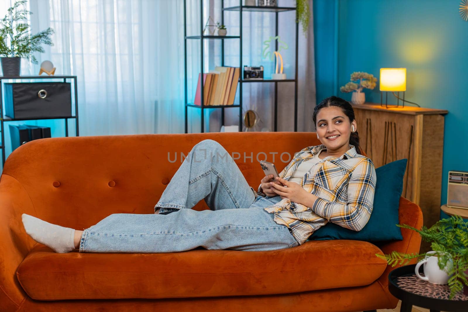 Indian young woman at home lying on sofa wearing earbuds, happily listening to music from smartphone. Arabian girl smiling, dancing slightly, relaxed atmosphere, enjoying pleasant musical moment