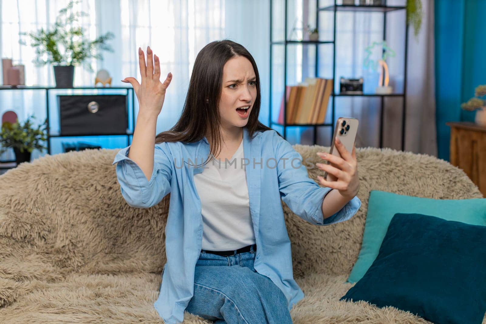 Young woman at home sitting on sofa irritated and emotionally arguing over video call, waving hand angrily. Caucasian girl expressing frustration, conflict, disagreement and stressed emotion. Quarrel
