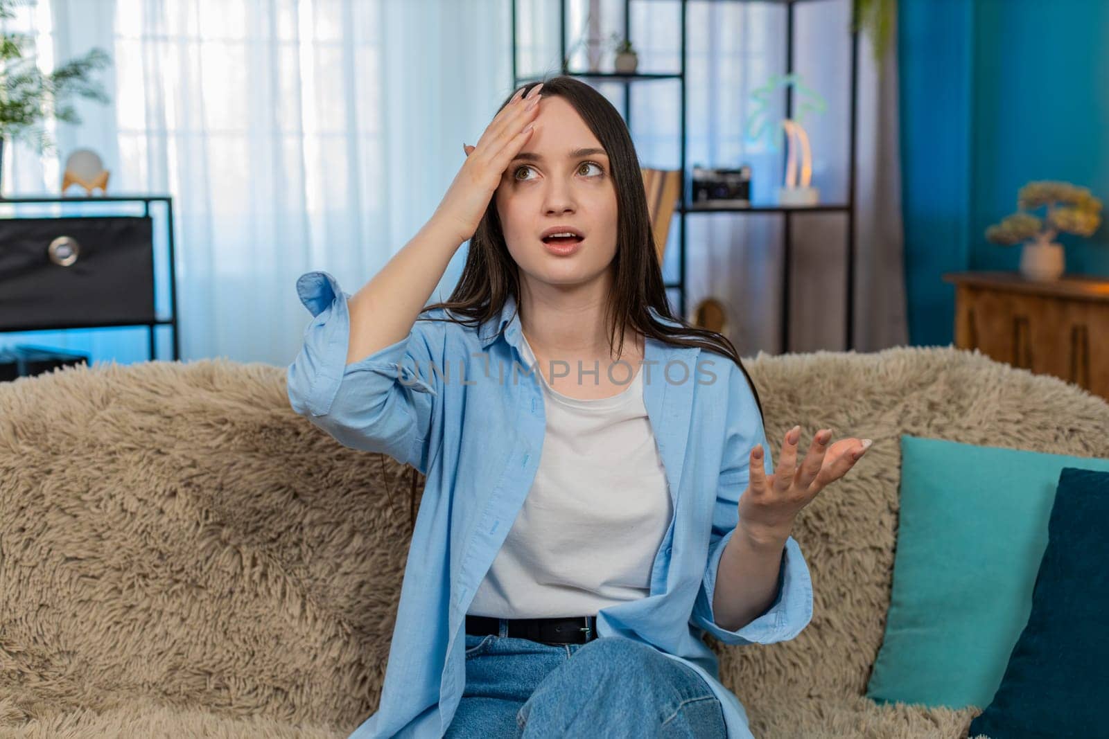 Young woman at home sitting on sofa upset and disappointed doing facepalm gesture, frustrated by her mistake. Caucasian girl demonstrating dissatisfaction, emotional regret and unhappy feelings.