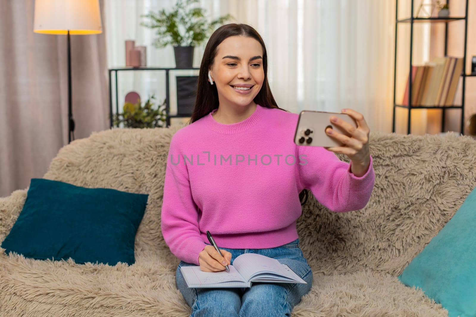 Young woman at home wearing headphones watching educational video lesson on mobile phone, taking notes attentively. Caucasian girl on sofa studying online, learning new information with interest