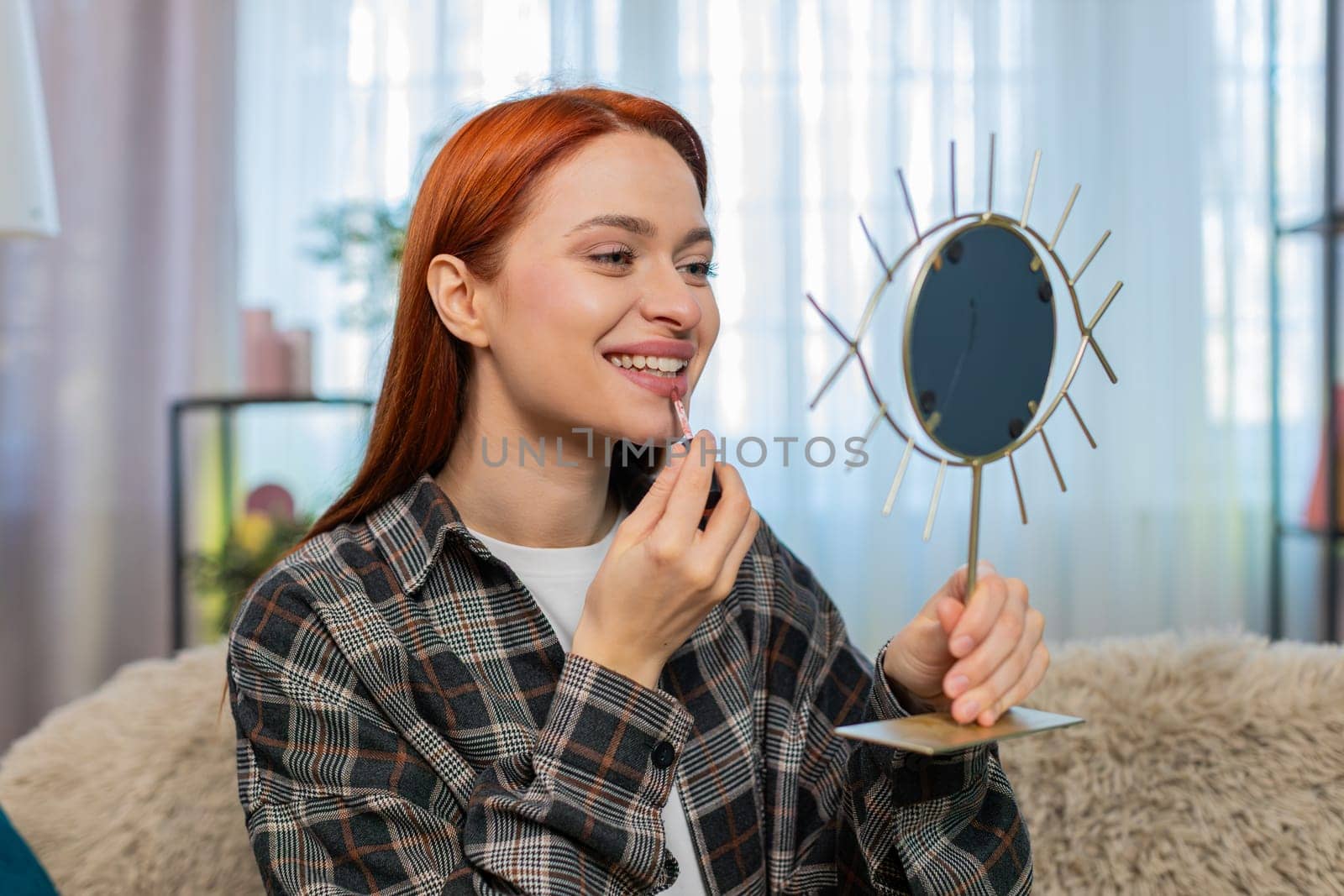 Young woman at home applying makeup with focus, drawing lips using lipstick and smiling into mirror. Caucasian redhead girl on sofa enjoying beauty routine with care, confidence and joyful emotion