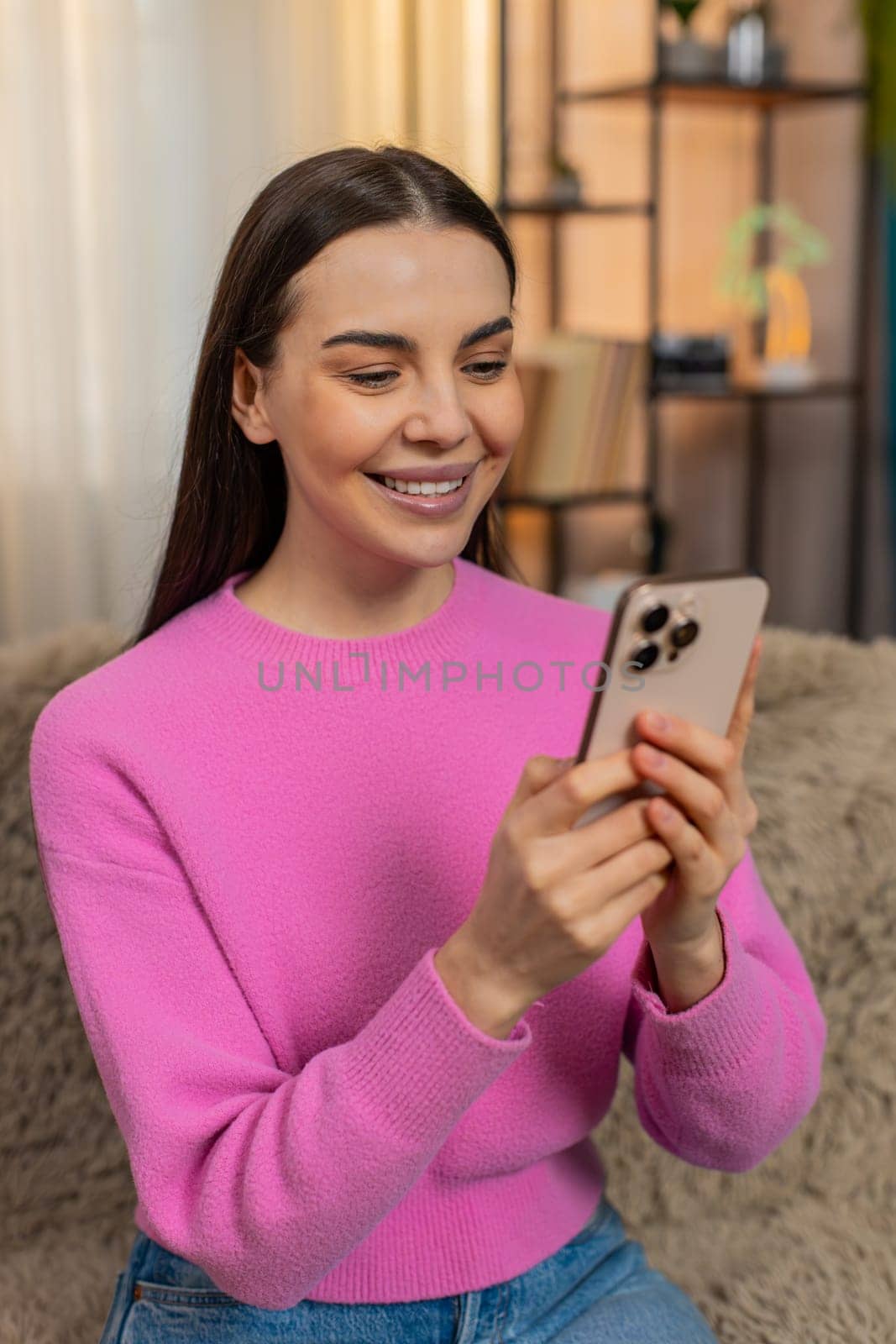 Young happy woman sitting at home, scrolling social media on mobile phone, suddenly smiling with joy and surprise. Caucasian girl relaxing on sofa, feeling amused, cheerful, happy, enjoying moment