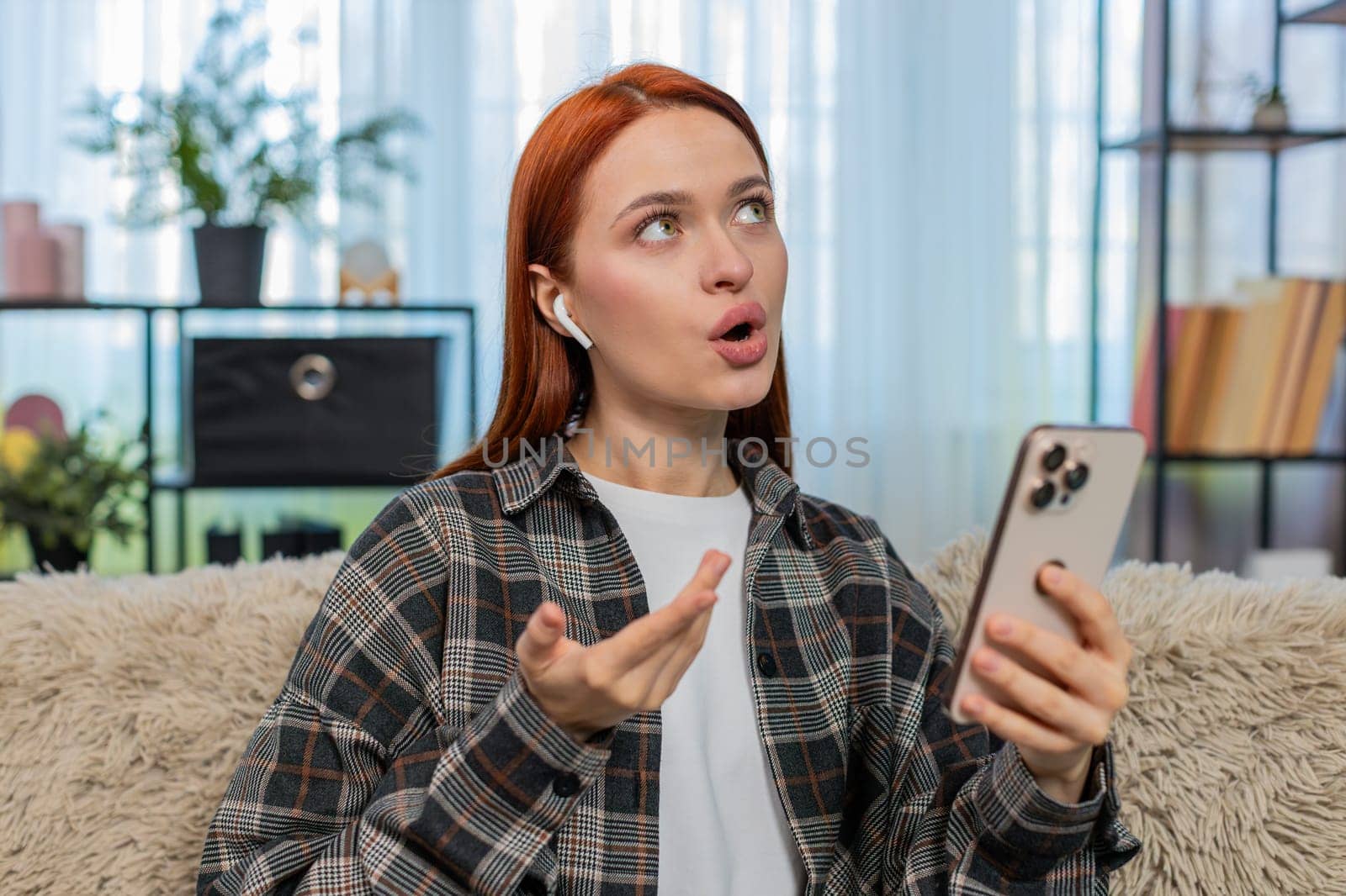 Young woman at home learning new language using mobile app and repeating words aloud with focus and joy. Caucasian redhead girl on sofa enjoying educational process practicing pronunciation and speech