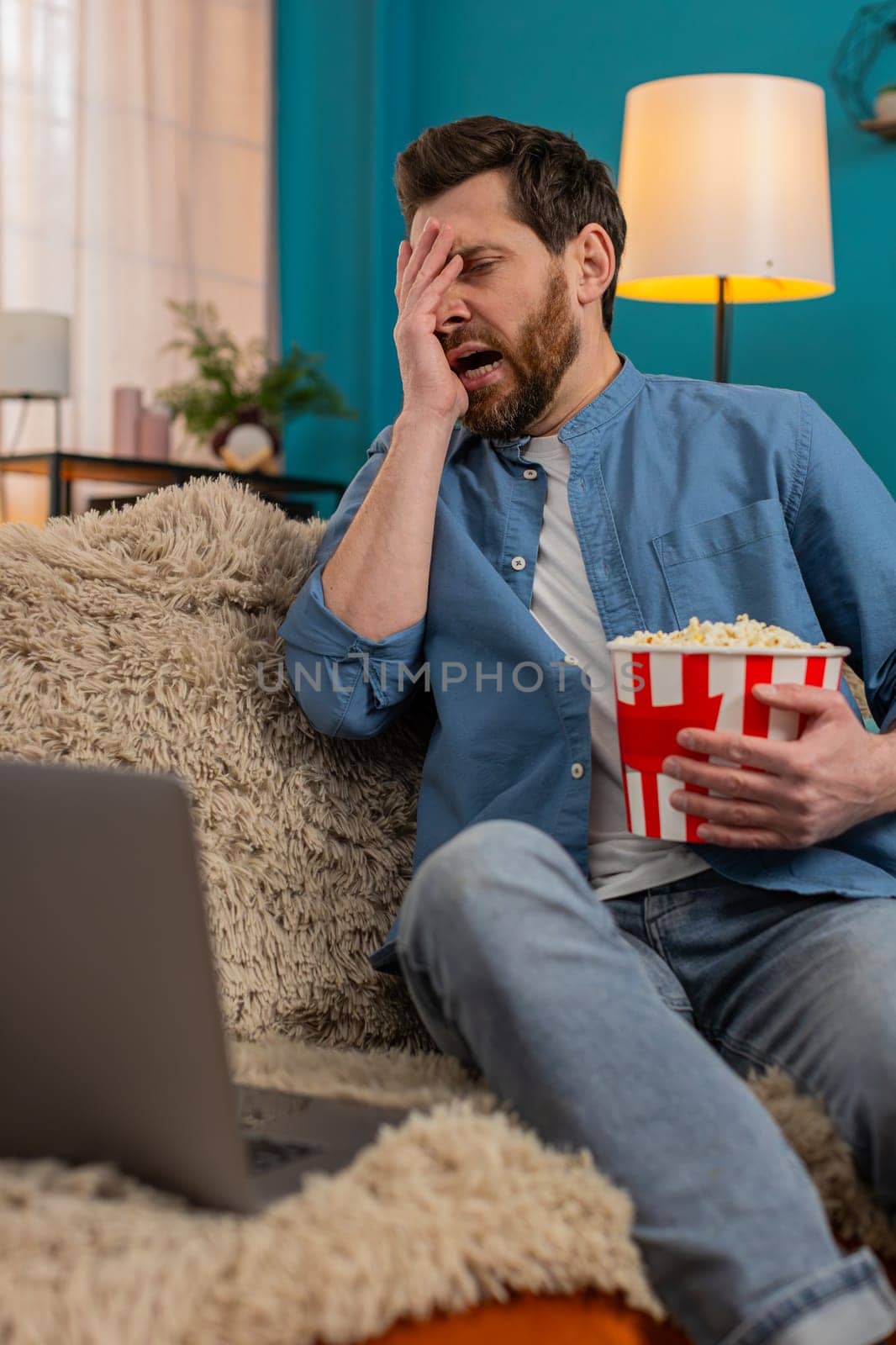 Adult man at home enjoys popcorn while watching movie on laptop, fully immersed in scene. Caucasian guy laughing in excitement at an unexpected moment, reacting emotionally to the thrilling storyline