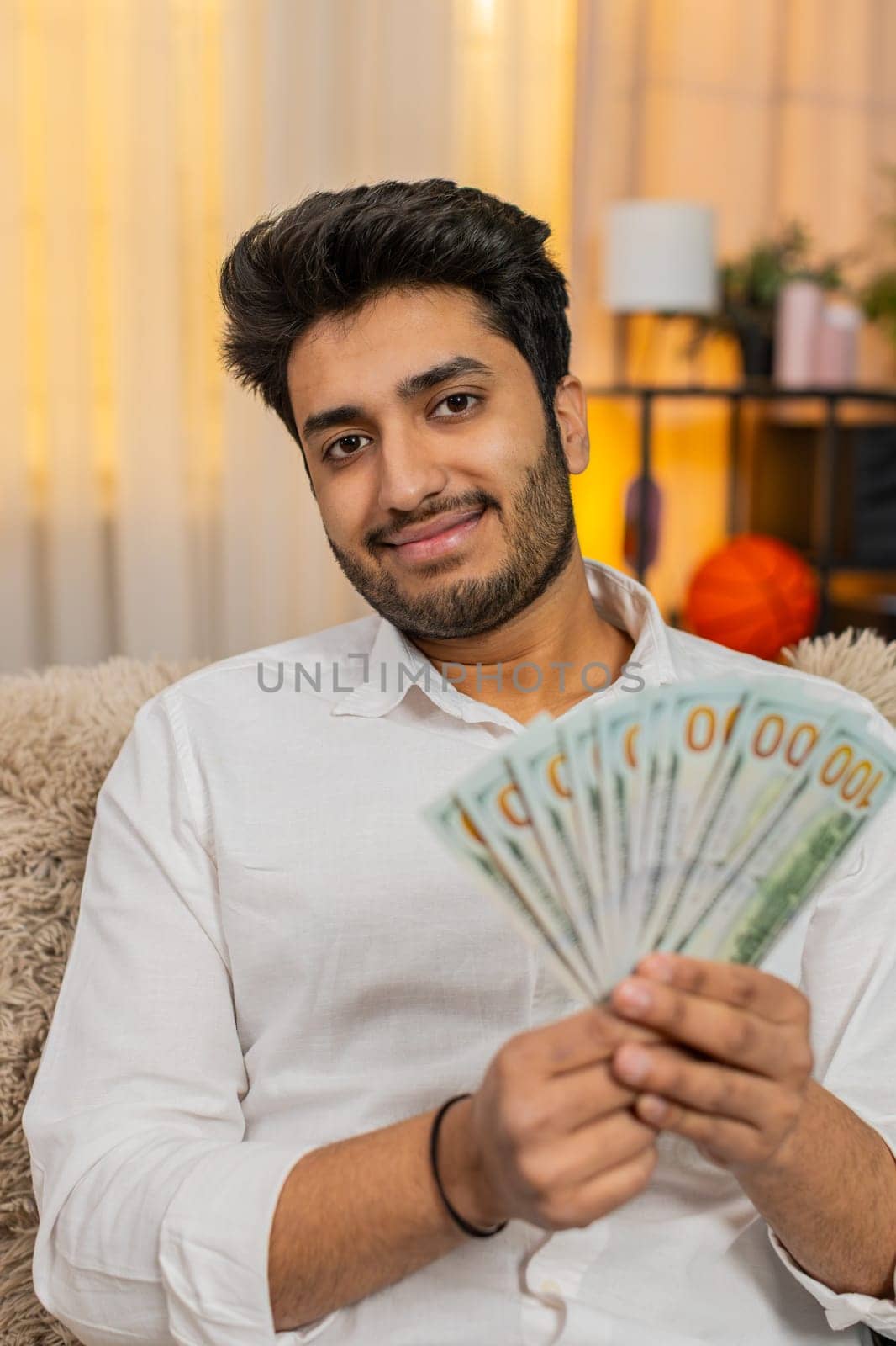 Indian man at home counting cash dollars, carefully considering his budget and planning future expenses. Arabian guy sitting on sofa, focusing on money management and thoughtful financial decisions