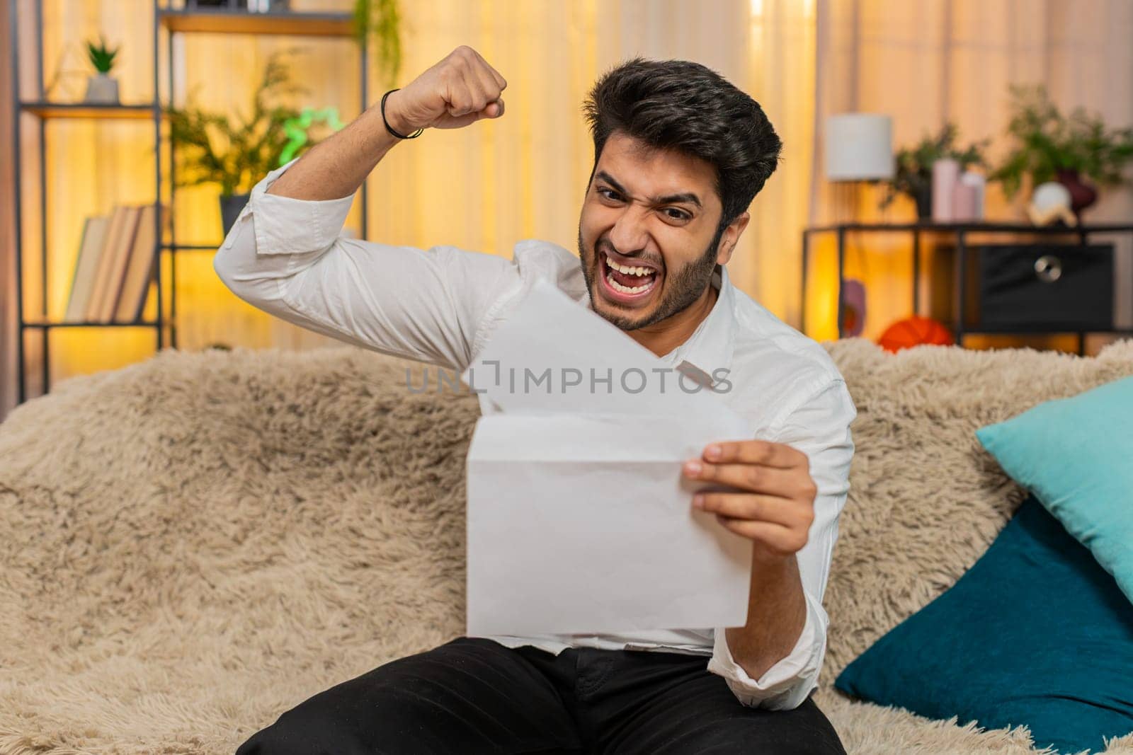 Indian man at home reading a letter with great news, celebrating a job offer, positive test results, or lottery win. Arabian guy expressing joy and excitement, celebrating success with a big smile