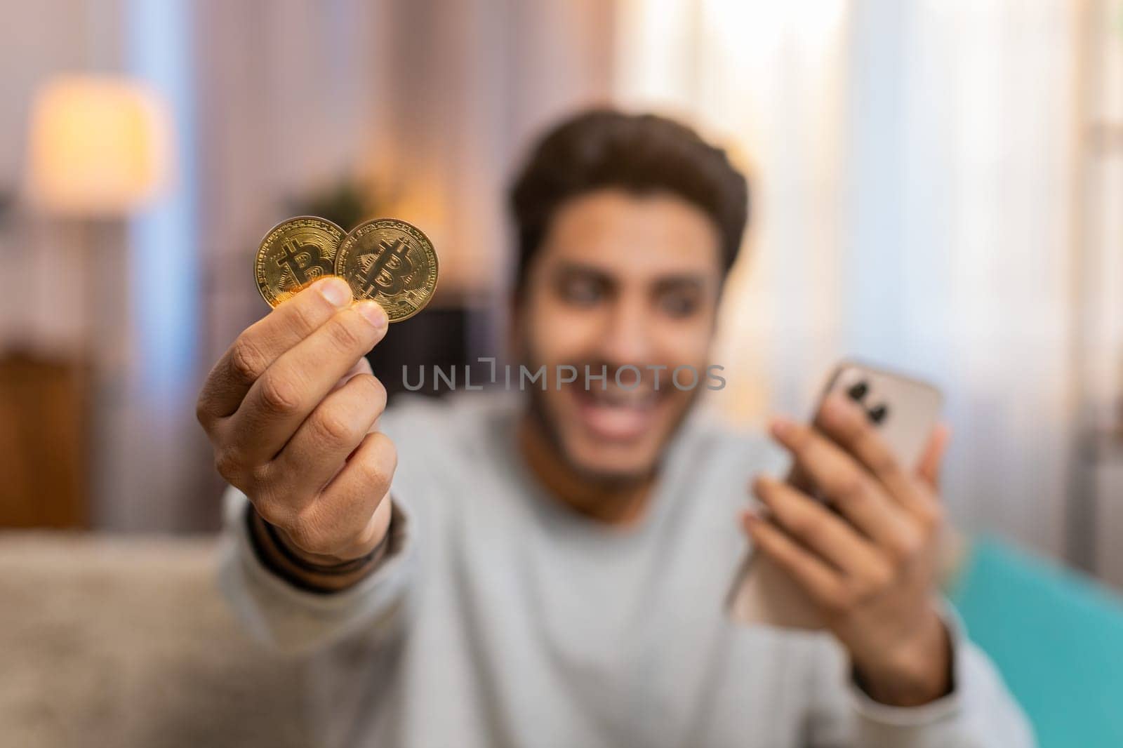 Young Indian man with bitcoin checks cryptocurrency exchange charts on smartphone app calculates approximate profits, looking on BTC coins in hand celebrating win. Hispanic guy looks satisfied at home