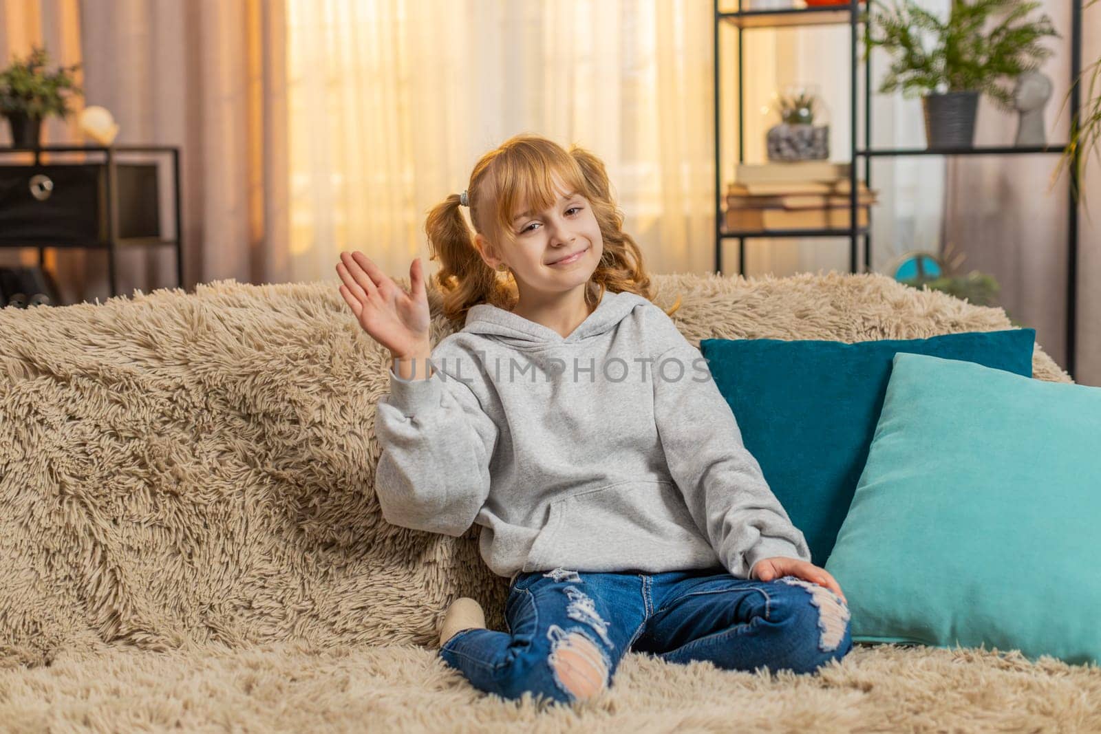 Young teenager girl waving hand with happy smile at home, expressing joy and friendliness. Caucasian child kid sitting on a sofa, saying hello hi welcome or goodbye with cheerful and relaxed attitude