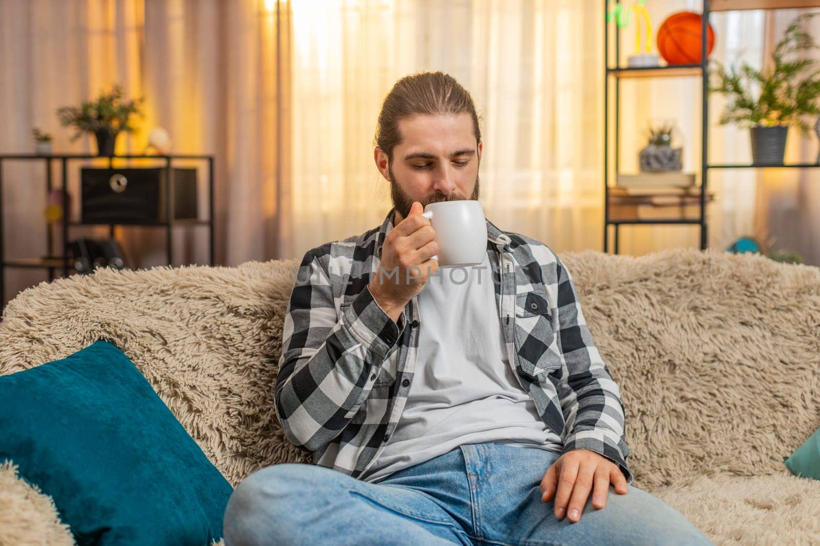 Young man holding steaming coffee or tea at home, closing eyes to soak smell in comforting fragrance. Guy on sofa inhales deeply, relishing each slow drinking sip mouthful with visible satisfaction.