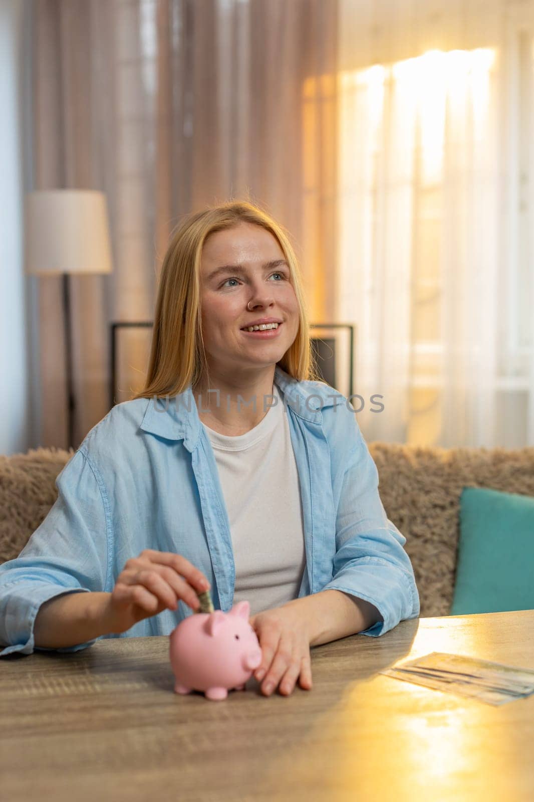 Young woman stores cash at home, dropping bills into piggy bank with contented grin and hopeful thought. Happy Caucasian girl sits at table radiates satisfaction aiming for future needs or investments