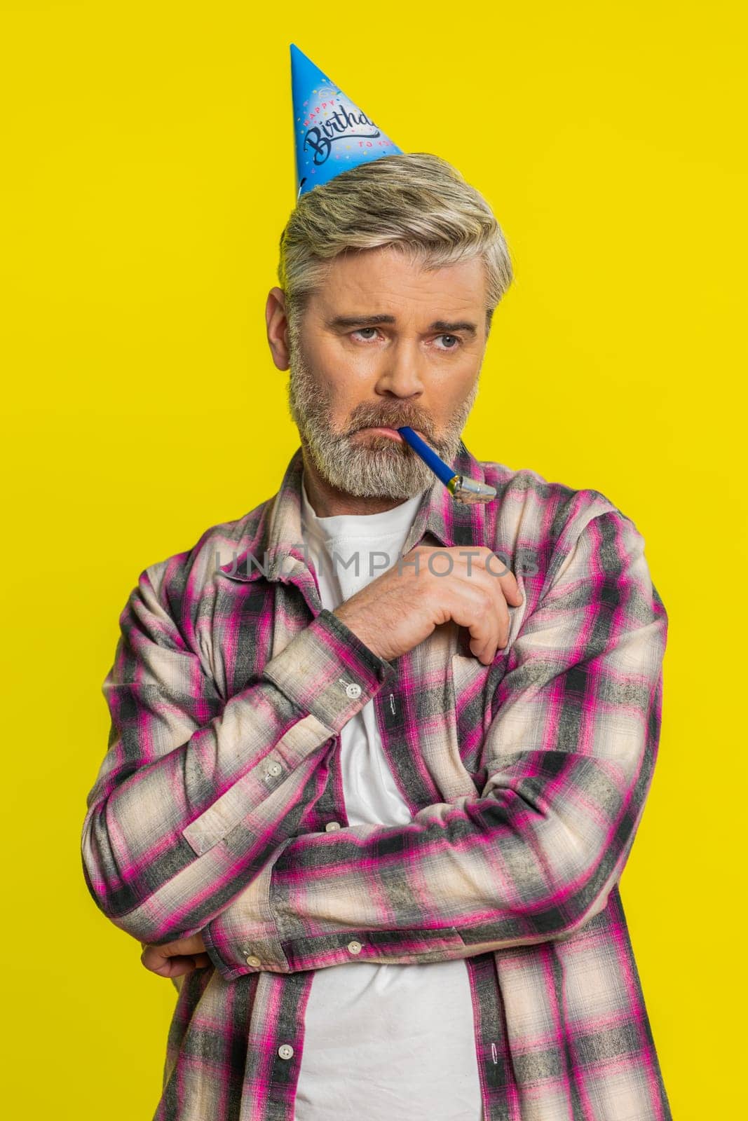 Mature man stands alone with party birthday cone hat and whistle blower showing dull spirit, disappointment. Middle-aged guy on yellow background feeling abandoned lonely, upset, tired, bored, unhappy