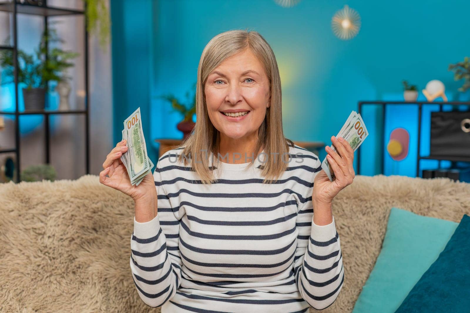 Happy Caucasian mature woman counts money dollar cash in hands after winning lottery bonus or receiving a gift. Grandmother smiles broadly sitting on home sofa, celebrating financial victory salary