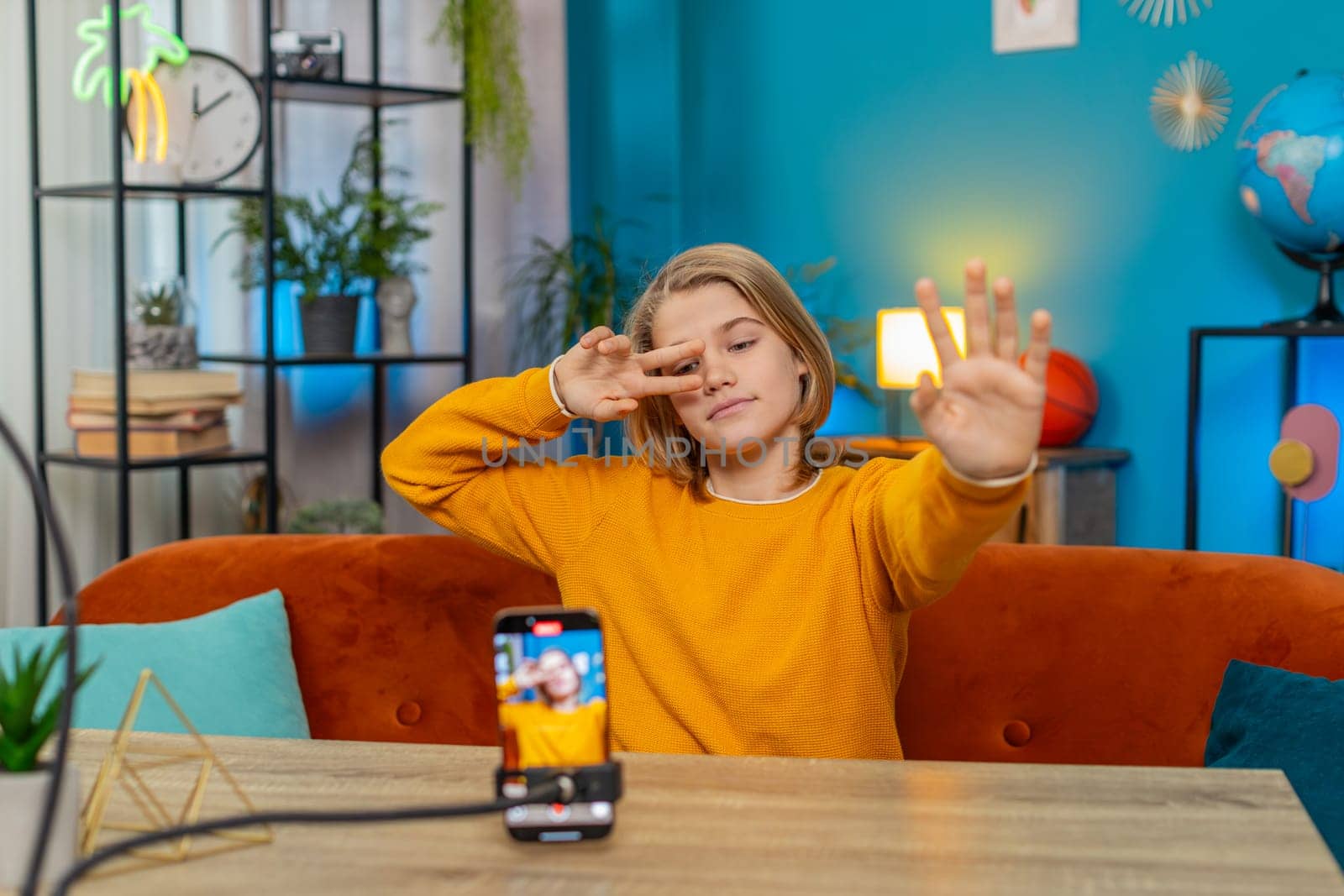 Caucasian teenager boy creating trendy selfie videos on smartphone at home. Performing dance moves while sitting on couch by table, engaging in social media content with enthusiasm and creativity