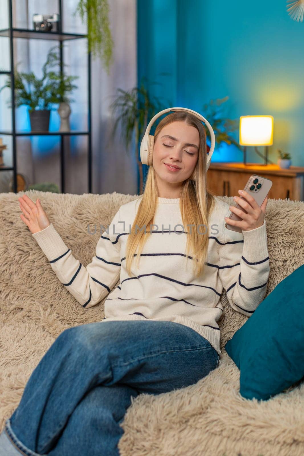 Caucasian happy young woman sitting on sofa at home, enjoying listening music with headphones, holding smartphone, closing eyes, smiling, feeling happy relaxed moving head to rhythm. Indoors lifestyle