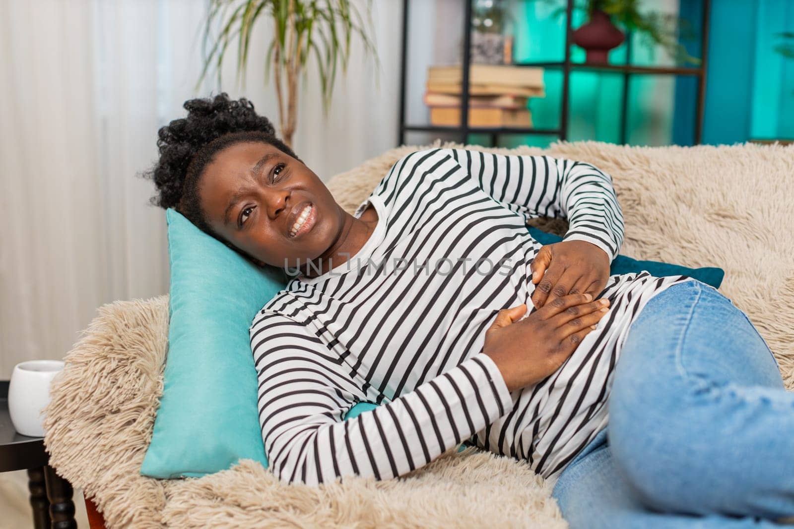 Black African American young woman suffering intense stomach pain, holding abdomen, lying on sofa at home. She looks distressed, tired, unwell, struggling with discomfort in cozy apartment living room