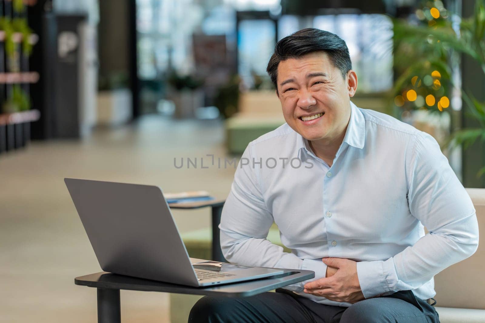 Disappointed sick Chinese businessman feeling overeating during work process on laptop computer in office lobby. Upset ill male manager guy suffering from painful stomach ache period cramps, poisoning