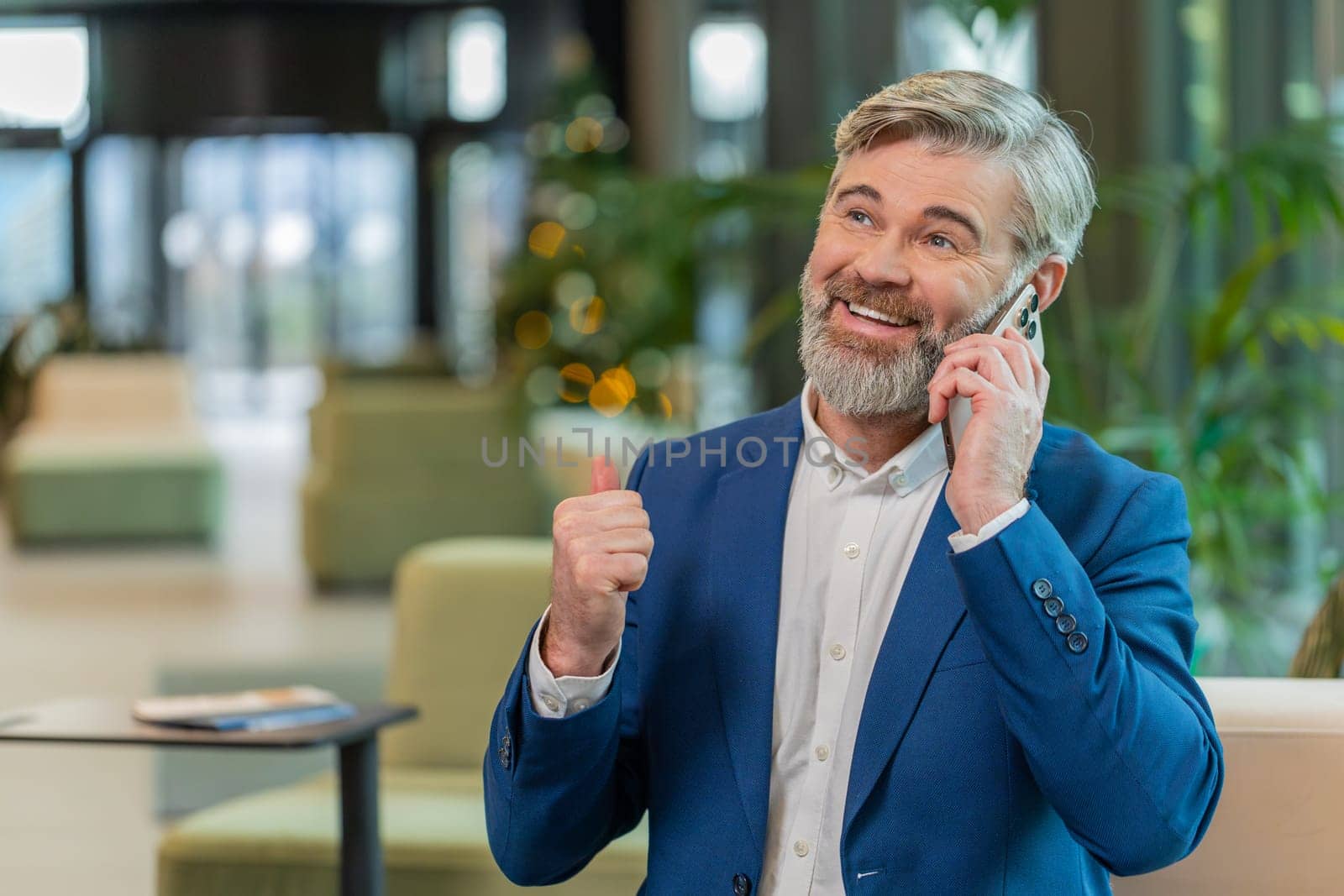 Happy mature businessman in suit answering to client on mobile phone call, using smartphone and laptop. Conversation. Caucasian bearded man guy working online job, talking in modern office lobby.
