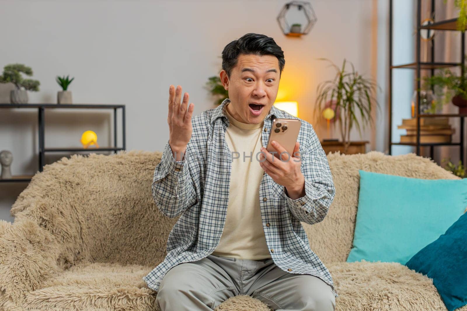 Asian middle-aged man sitting on sofa at home reading unexpected positive email news on smartphone. Surprised, celebrating online game victory. Happy, joyful man indoors enjoying successful moment