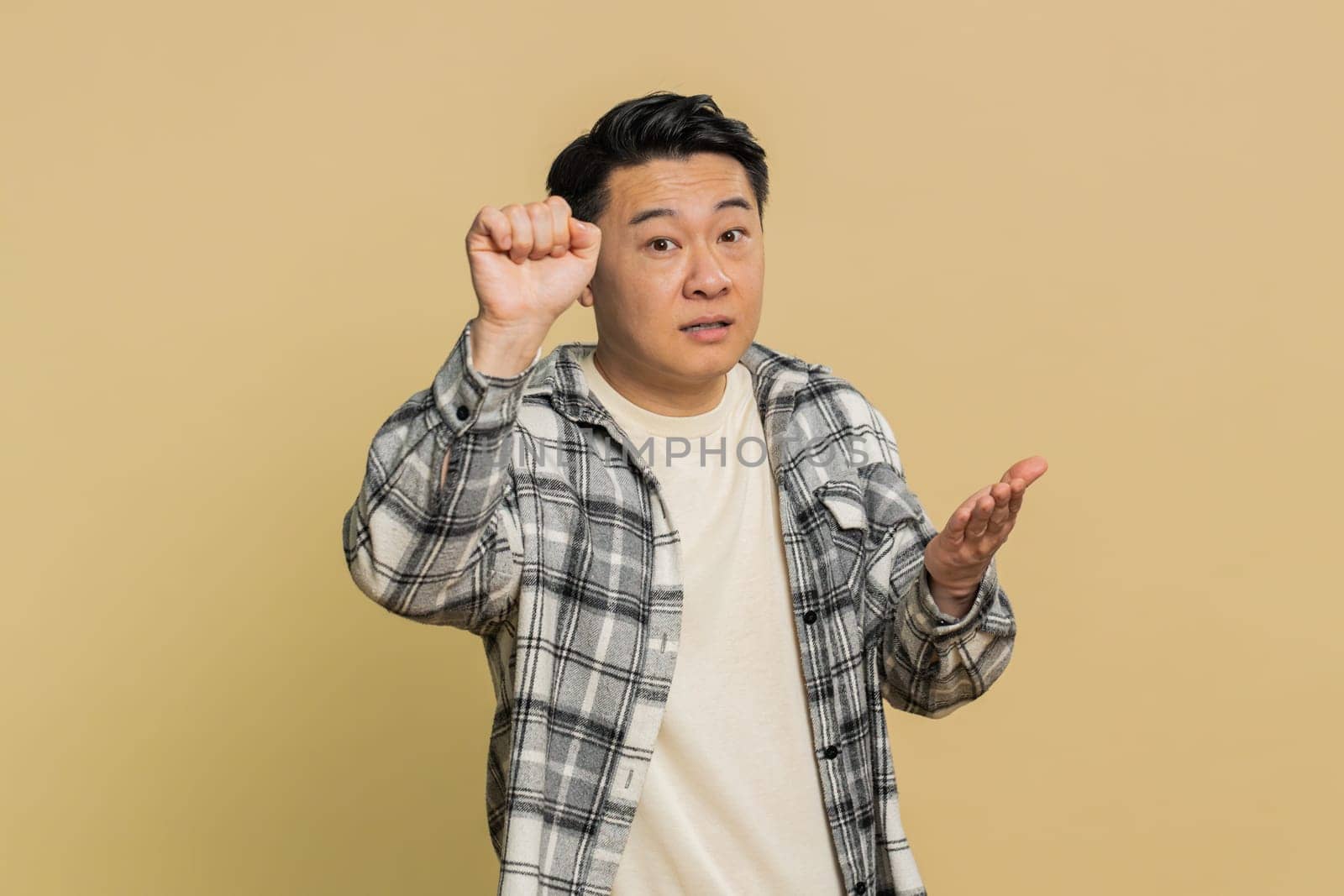 Knock-knock who is there. Confused excited Asian man knocking door gesture asking who is at home, feeling embarrassed, no idea, being clueless and uncertain. Mature Chinese guy on beige background