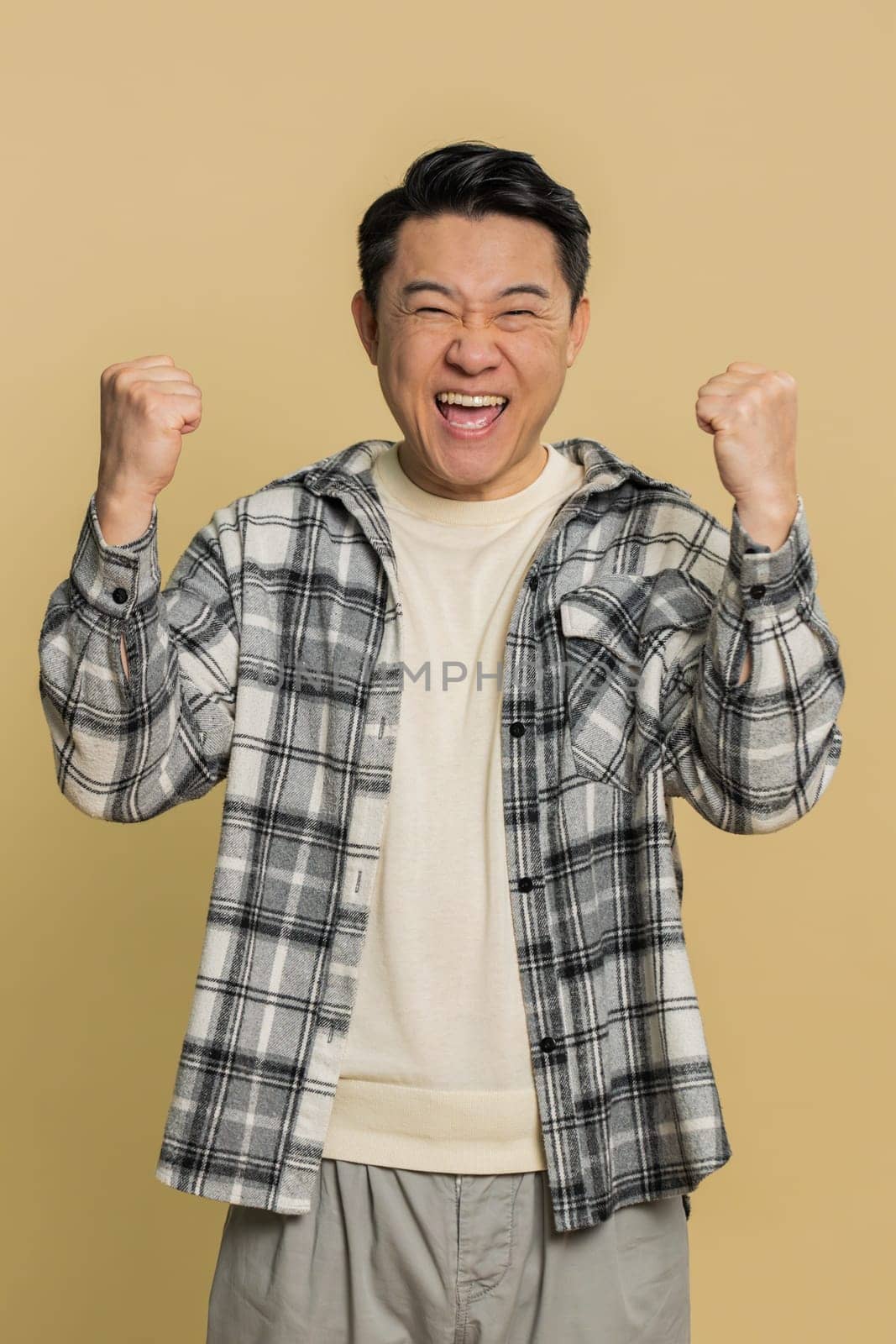 Asian middle-aged man celebrating success victory, winning birthday, lottery jackpot goal achievement, play game, good positive news, triumph. Mature adult guy isolated on beige background indoors