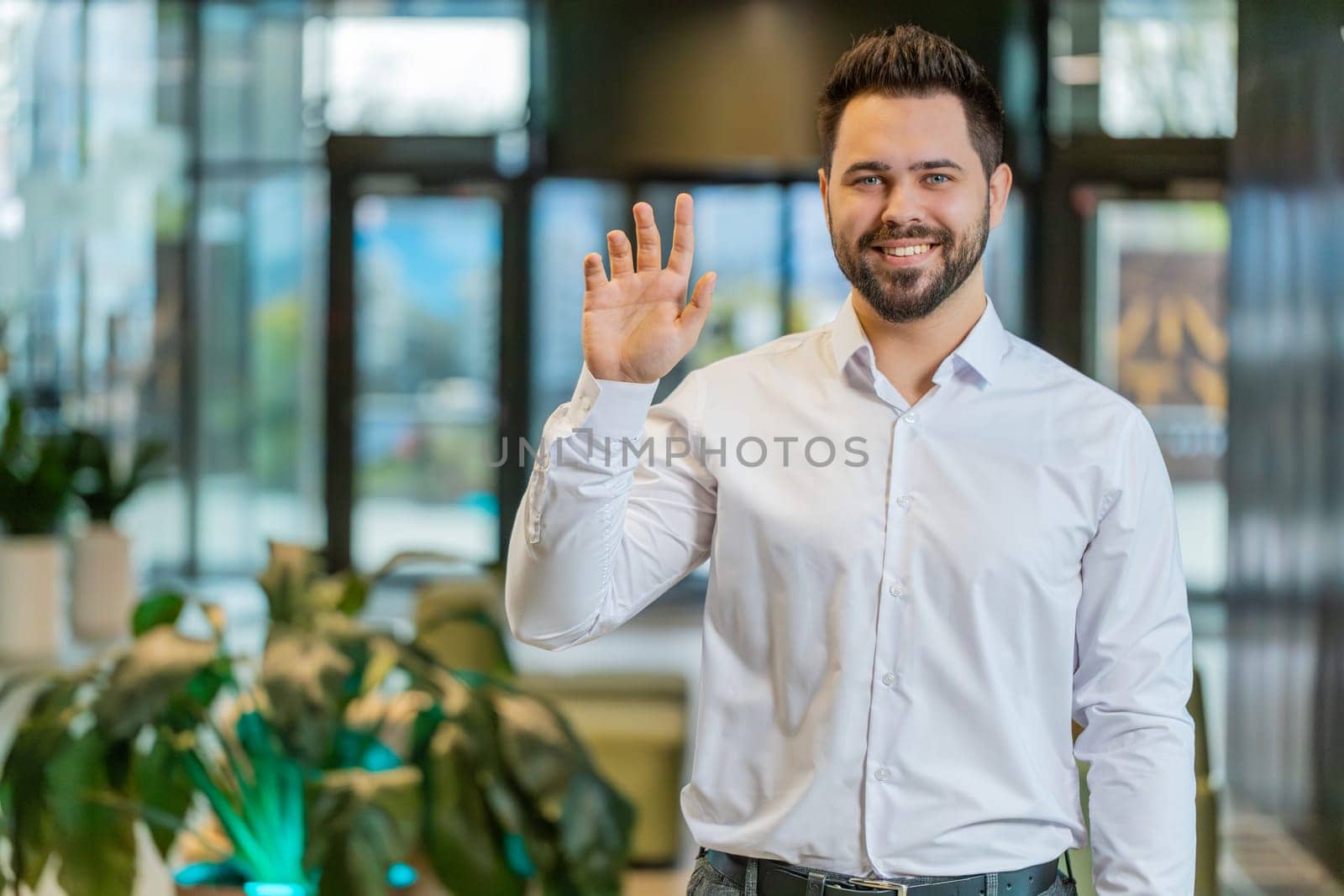 Hello. Happy young Caucasian businessman employee waves hand hi gesture greeting welcomes someone, online webinar, remote distance conversation. Entrepreneur man in shirt standing in office lobby.